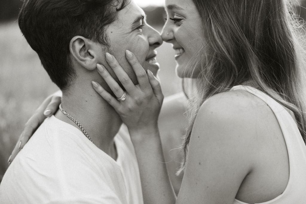 A close-up black and white photo of a couple nearly kissing, her engagement ring prominently visible as she holds his face tenderly