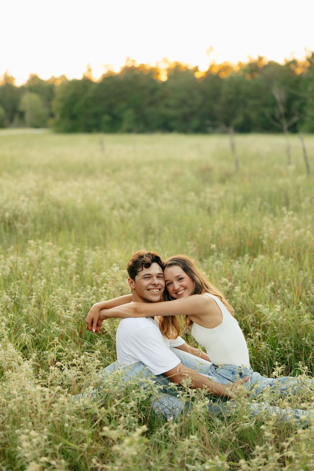 A couple sits side by side in a wildflower meadow at golden hour, laughing together with their heads touching, both in white tops and light denim