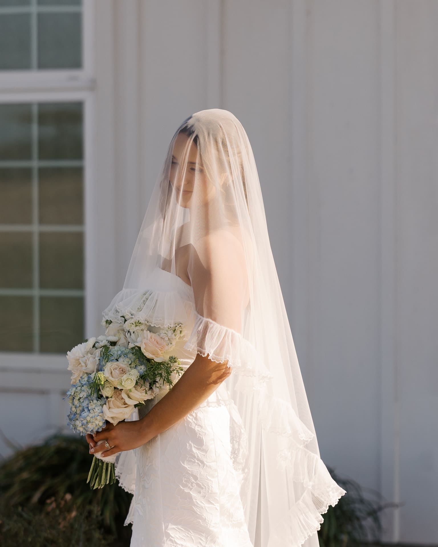 Soft bridal portraits of a bride in an off-the-shoulder lace gown, veil draped over her face as she holds a lush bouquet of blush roses and blue hydrangeas in warm golden light