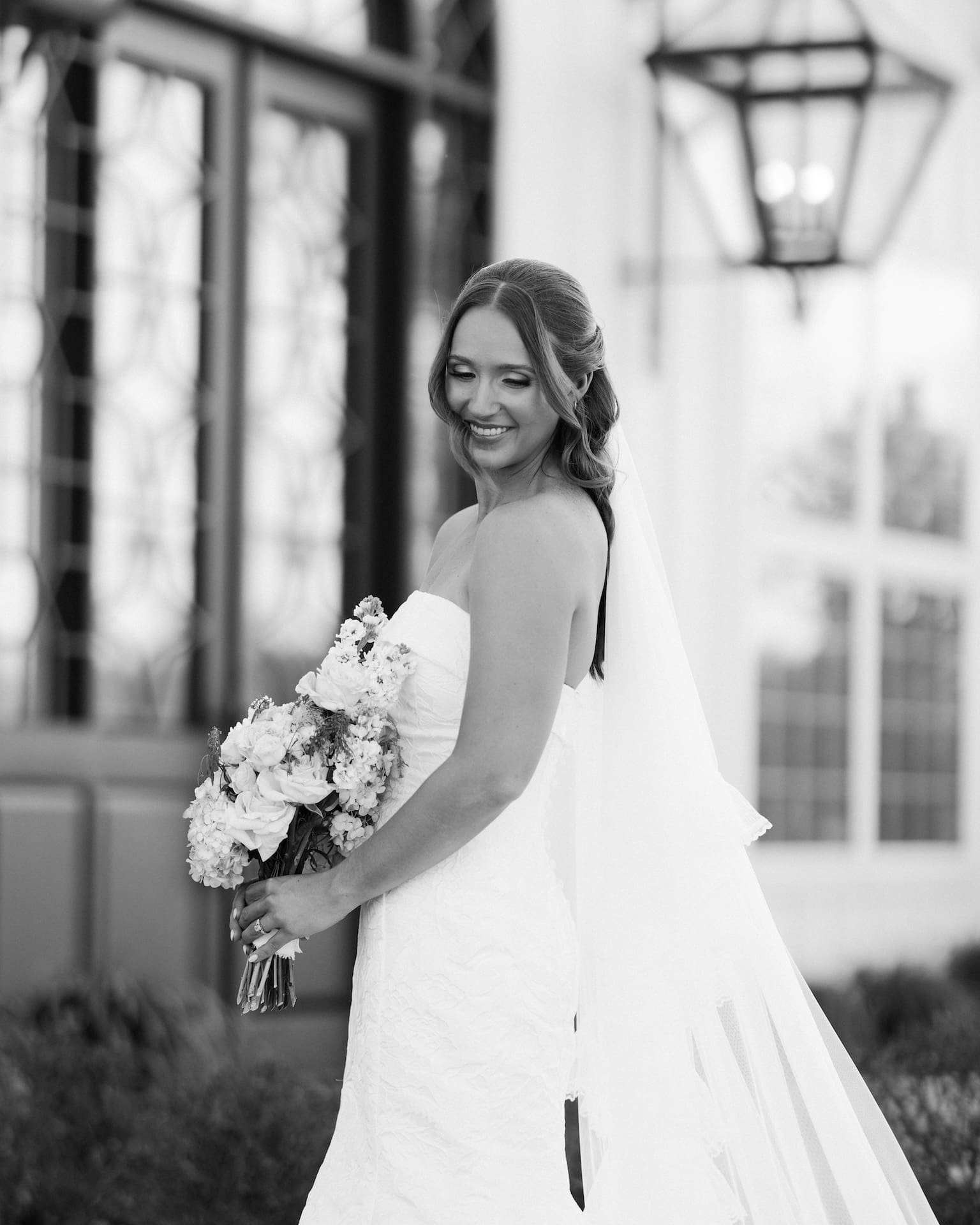 Black and white bridal portraits of a bride in a strapless gown glancing back with a soft smile, her veil cascading down as she stands in front of glowing iron lanterns at a white barn venue