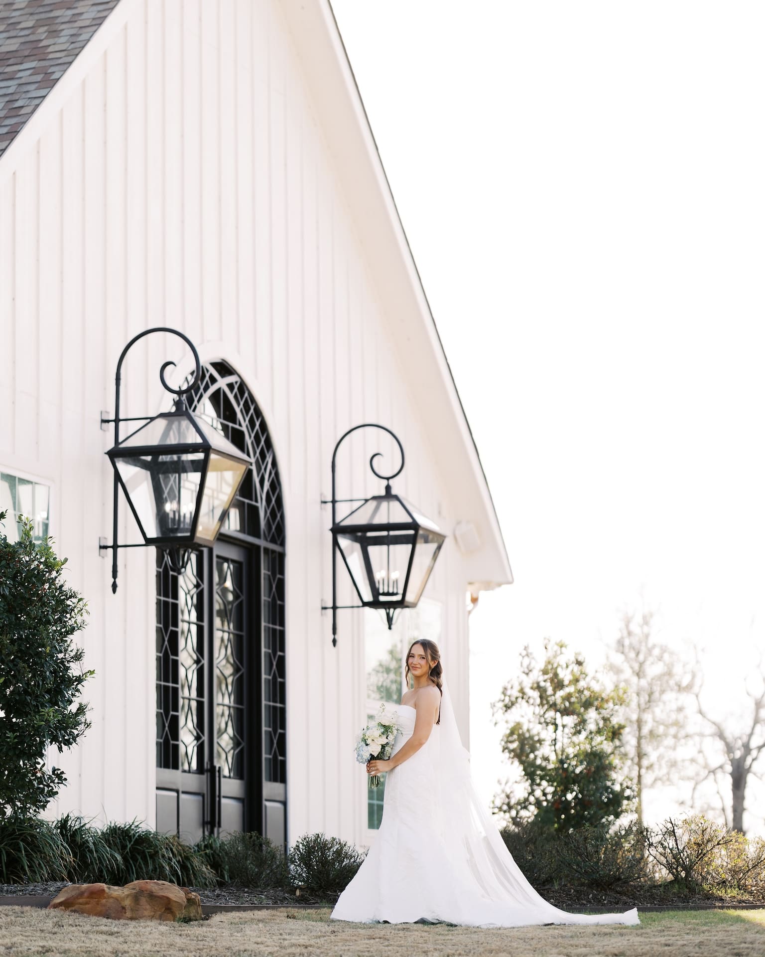 Wide-angle bridal portraits of a bride in a strapless mermaid gown smiling beside the arched iron-paned entrance of a white barn chapel, flanked by two large black lanterns at golden hour