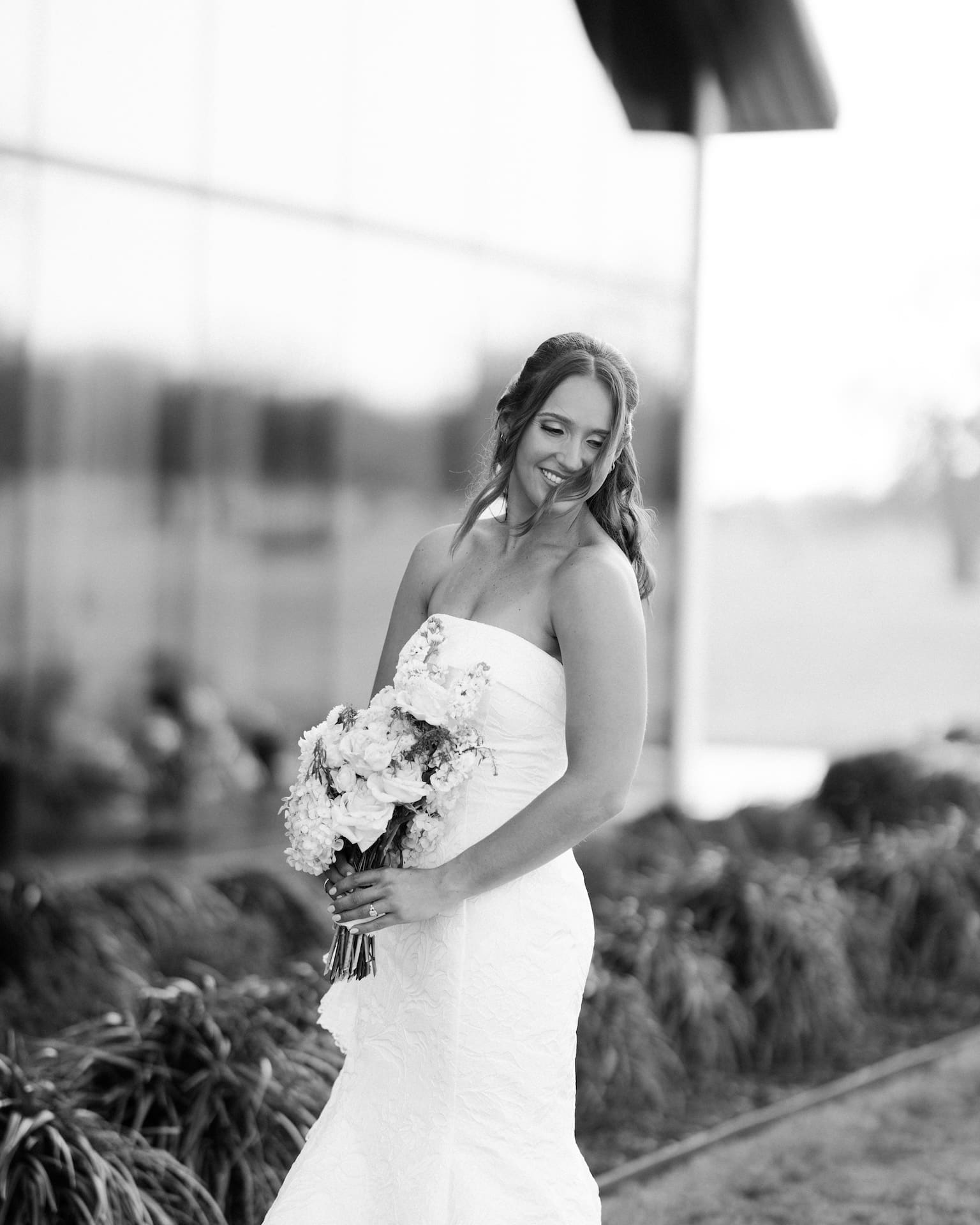 Candid bridal portraits of a bride in a strapless lace gown laughing and looking down as she holds a white bouquet outside a glass-walled venue
