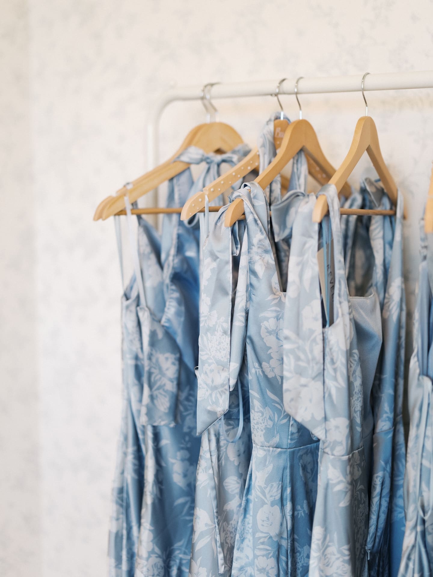 The bridesmaids' dusty blue floral satin gowns hanging in a row on wooden hangers, ready for the wedding day.