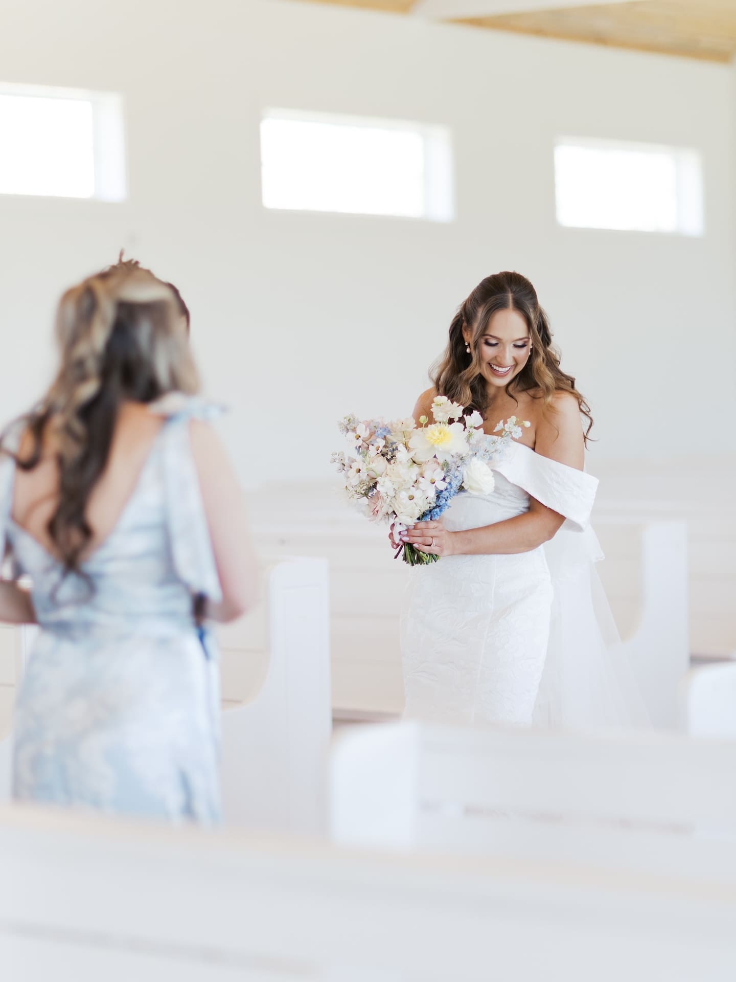 The bride laughs down at her bouquet during a sweet moment with a bridesmaid inside the bright white chapel.