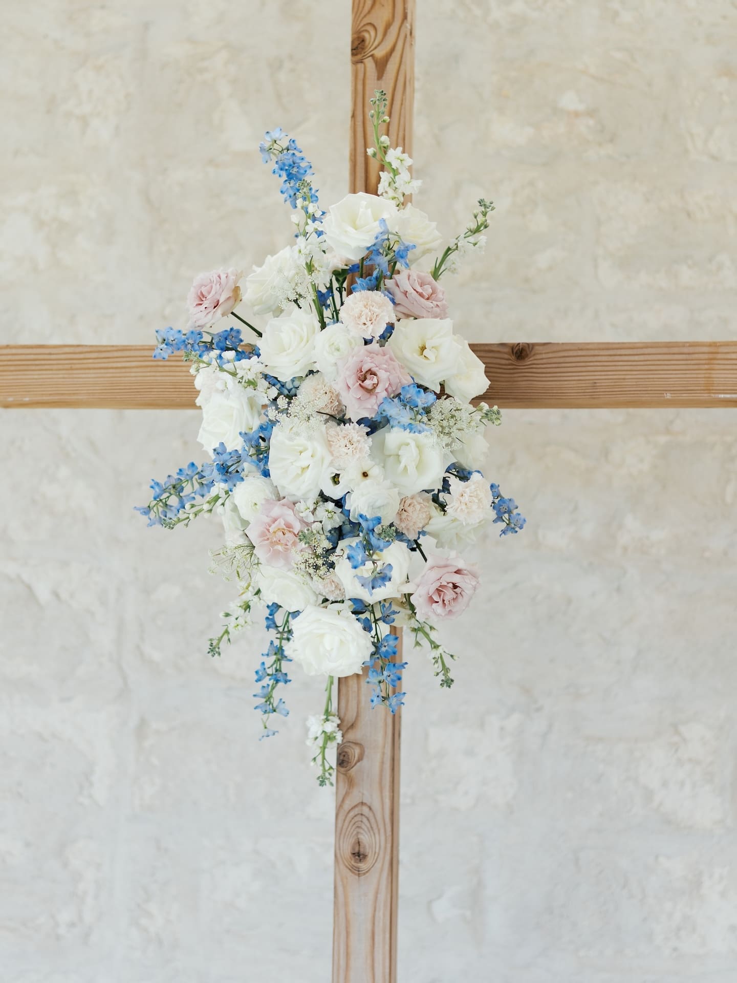 A close-up of the wooden altar cross adorned with blush roses, white lisianthus, and blue delphinium blooms for the ceremony.
