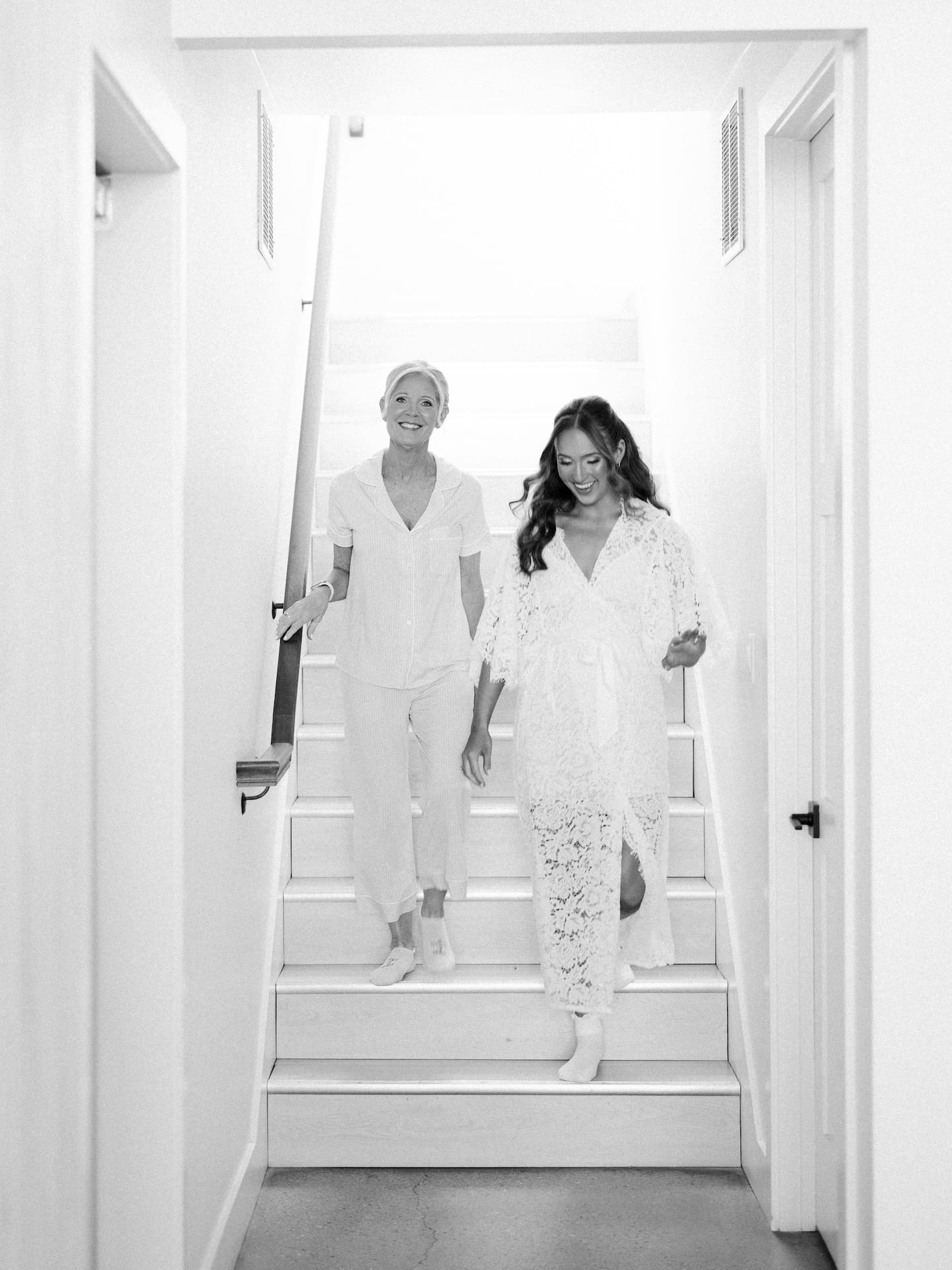 A black and white film photo of the bride and her mom walking down the stairs together on the morning of the wedding, both smiling and glowing.