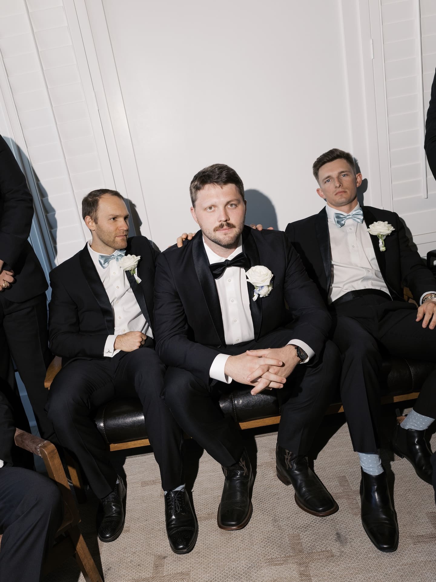 
The groom sits confidently between two groomsmen at Camp Hosea, all three in classic black tuxedos with white boutonnieres, just before the ceremony.