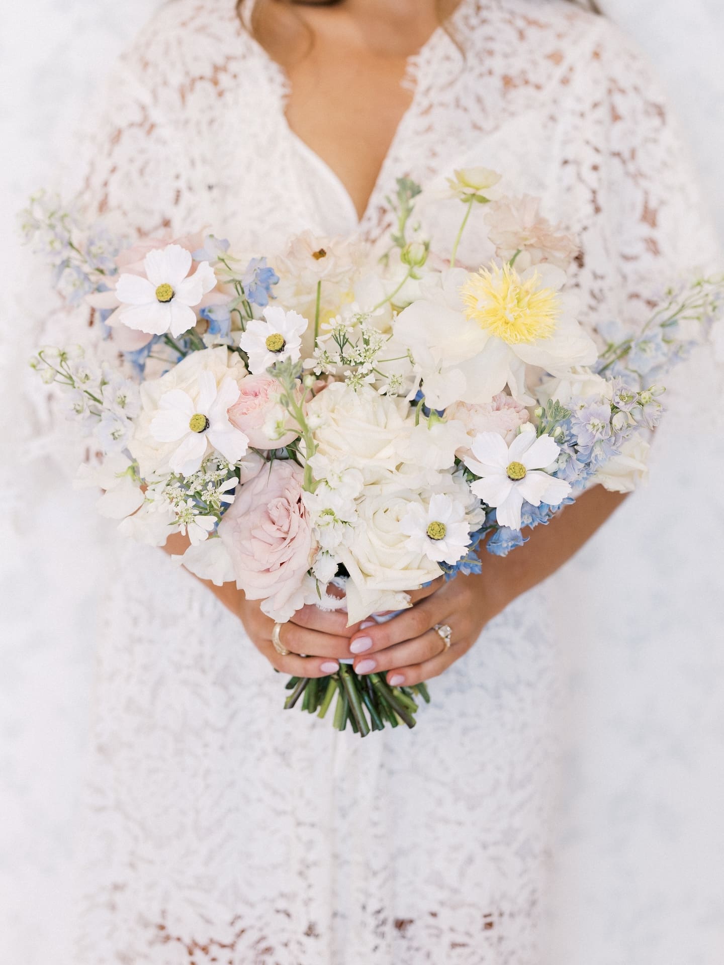 The bride holds her lush garden-style bouquet of white cosmos, blush roses, and blue delphinium at Camp Hosea.