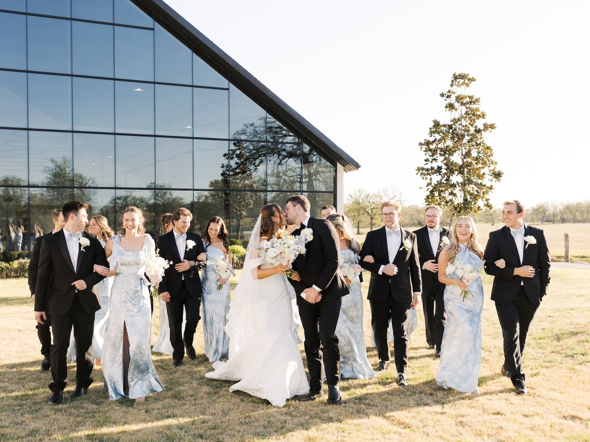 The wedding party walks and laughs together across the grounds at Camp Hosea, the bride and groom stealing a kiss at the center of it all.
