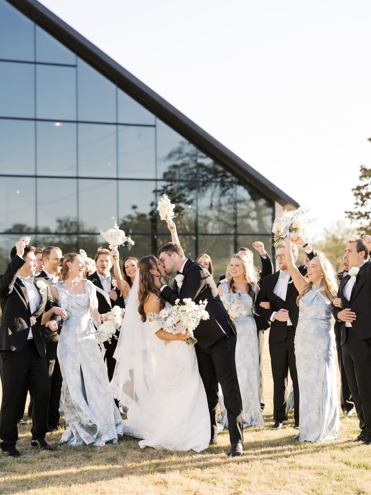 The entire wedding party erupts in cheers in front of the Camp Hosea glass chapel as the bride and groom share a kiss, bouquets and fists raised in celebration.