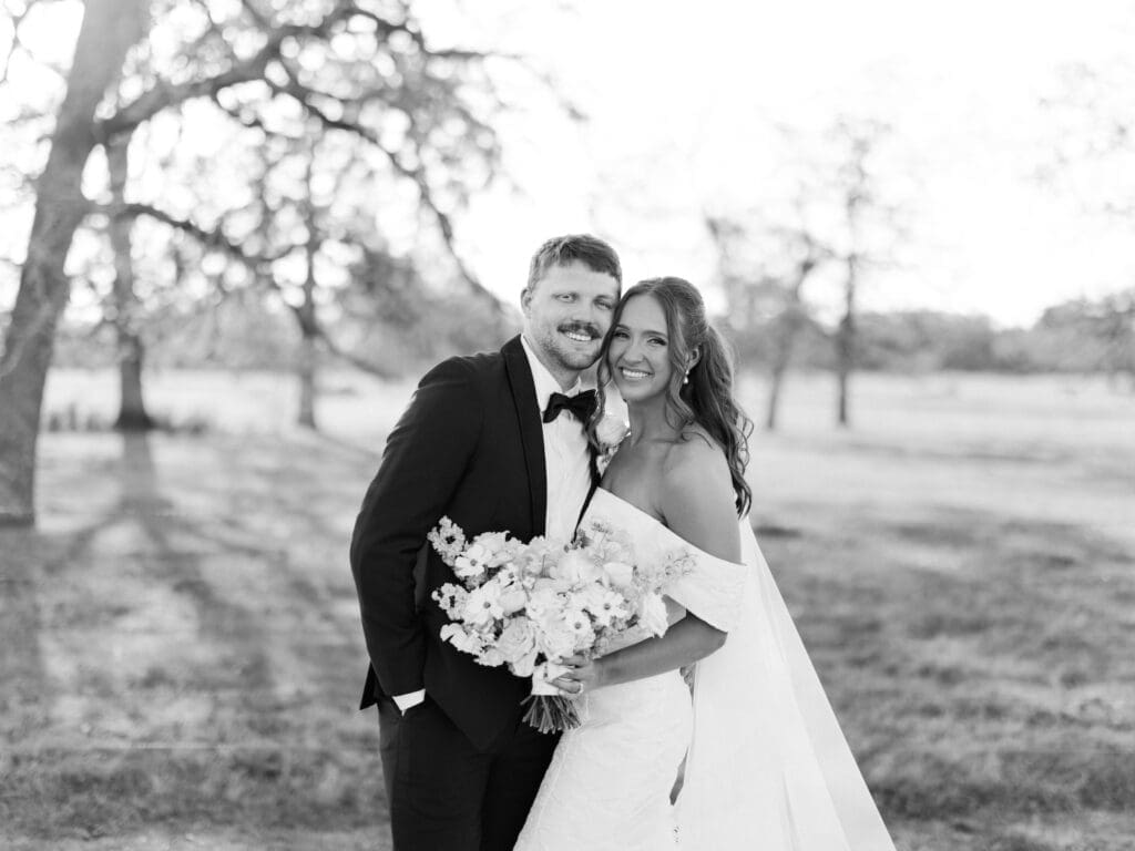A classic black and white film portrait of the bride and groom smiling together on the open grounds at Camp Hosea, bare trees softly blurred behind them.