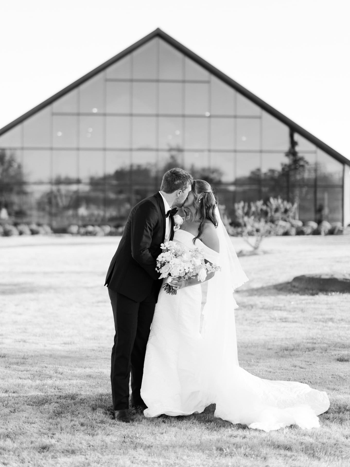 A timeless black and white portrait of the bride and groom kissing on the lawn in front of the iconic glass chapel at Camp Hosea.