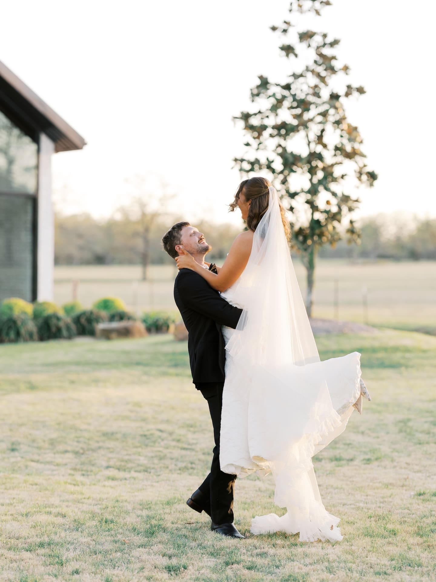 The groom lifts the bride up on the sun-drenched lawn at Camp Hosea, both of them laughing as her veil and skirt float in the air.