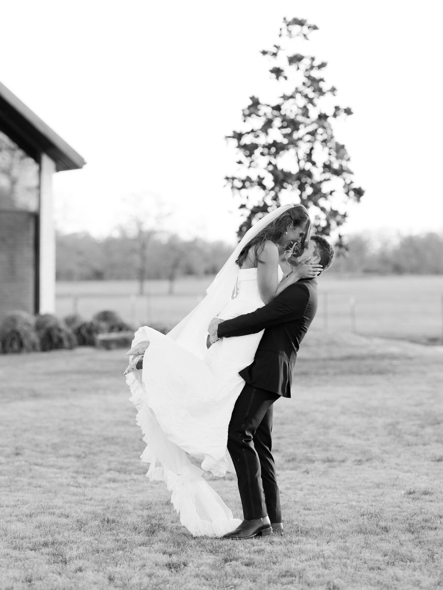 A black and white film photo of the groom lifting the bride for a kiss on the lawn at Camp Hosea, her gown and veil sweeping dramatically around them.