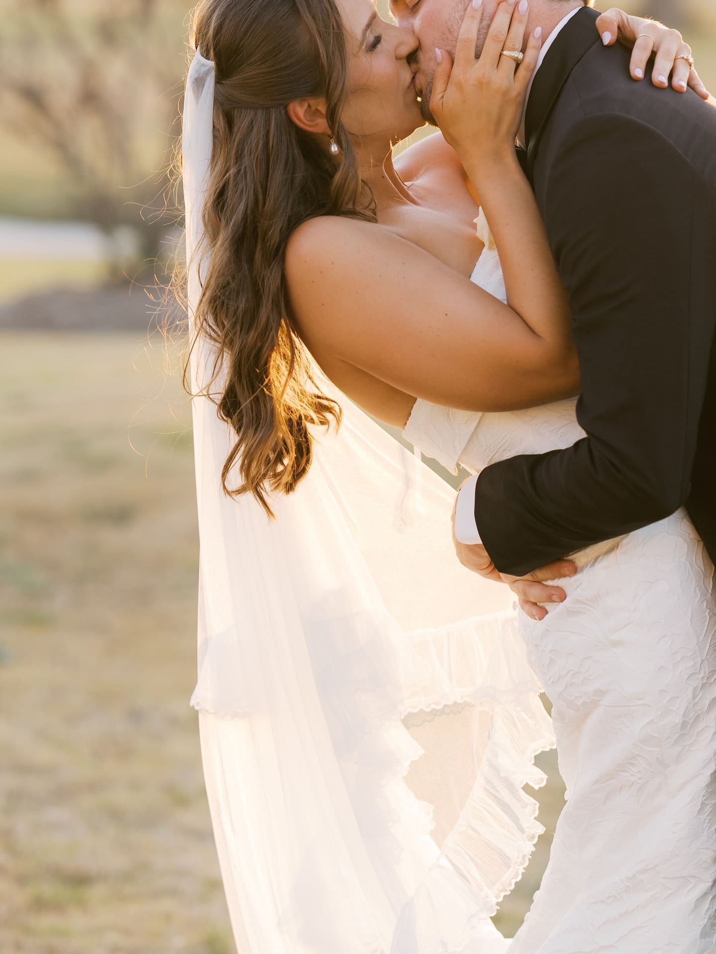 The bride and groom share a golden hour kiss on the grounds of Camp Hosea, warm light catching her veil and the delicate lace of her gown.