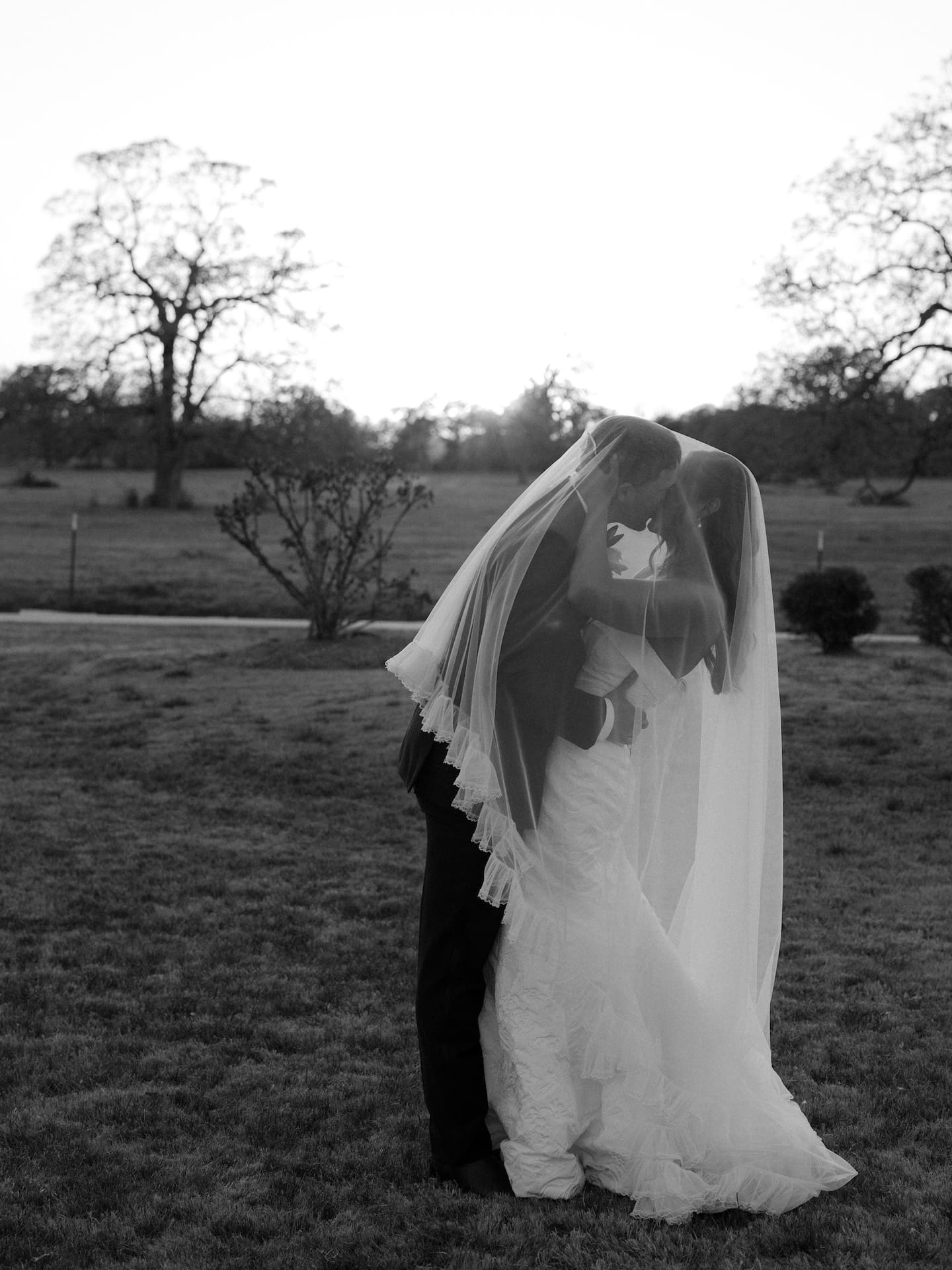 A stunning black and white film portrait of the bride and groom kissing beneath her veil on the open lawn at dusk, bare winter trees silhouetted behind them.