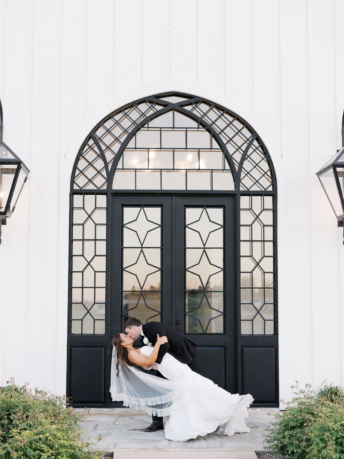 The groom dips the bride for a kiss in front of the stunning arched iron doors of the Camp Hosea chapel, lanterns glowing on either side of them.
