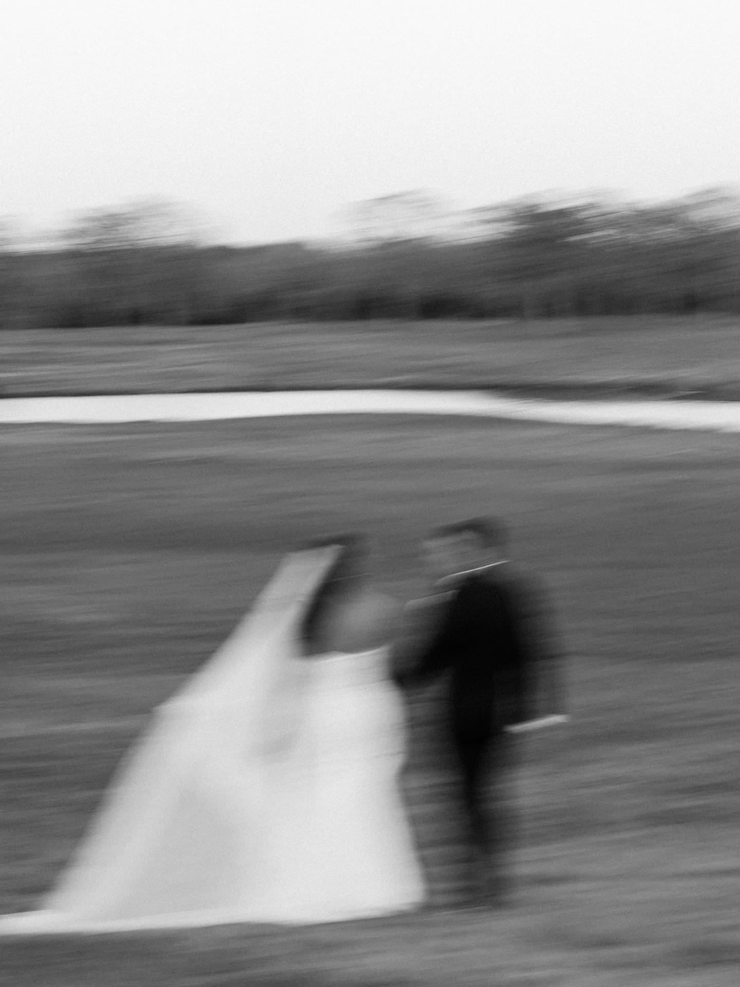 A dreamy, motion-blurred black and white film photo of the bride and groom walking hand in hand through an open field.