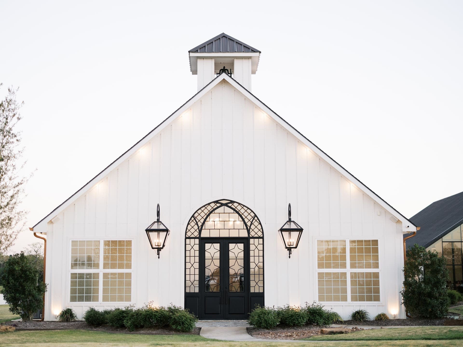 The exterior of the Camp Hosea chapel glows warmly at dusk, its white board-and-batten facade, arched iron doors, and lantern sconces lit against the evening sky.