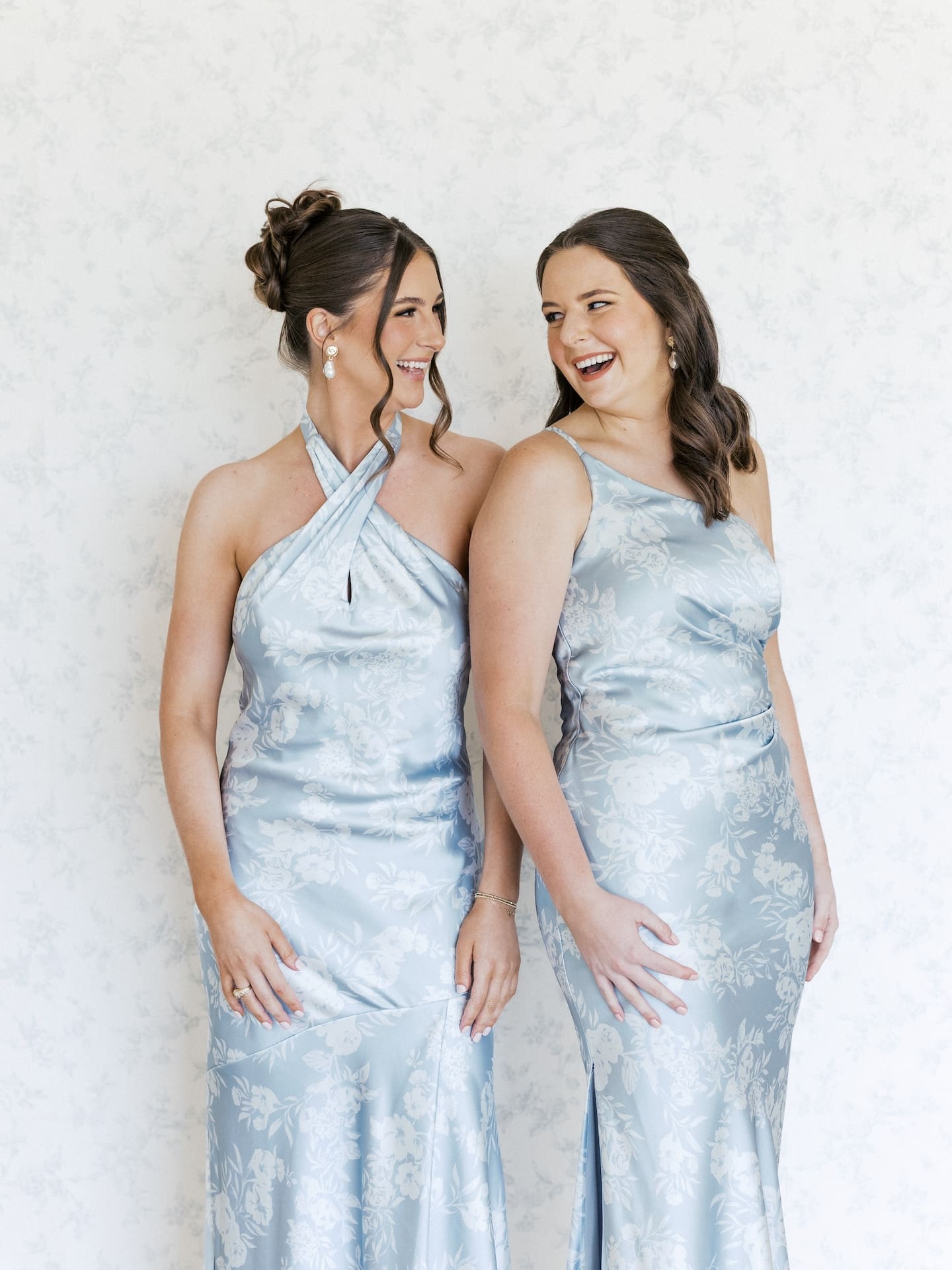 Two bridesmaids in dusty blue floral gowns laugh together, caught in a genuinely joyful moment during getting-ready portraits.