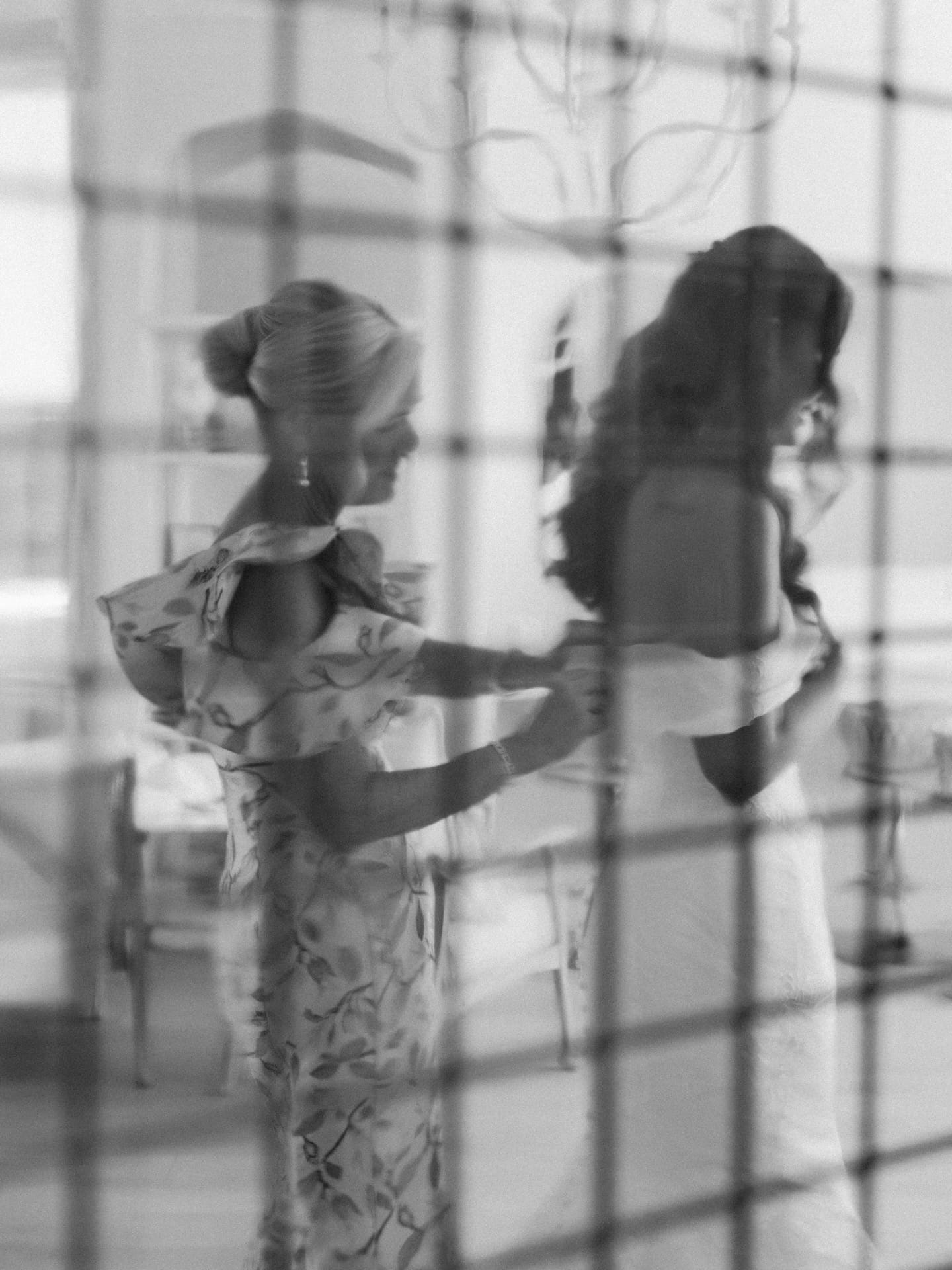 An artistic black and white image shot through a gridded glass pane of a bridesmaid helping the bride get dressed.