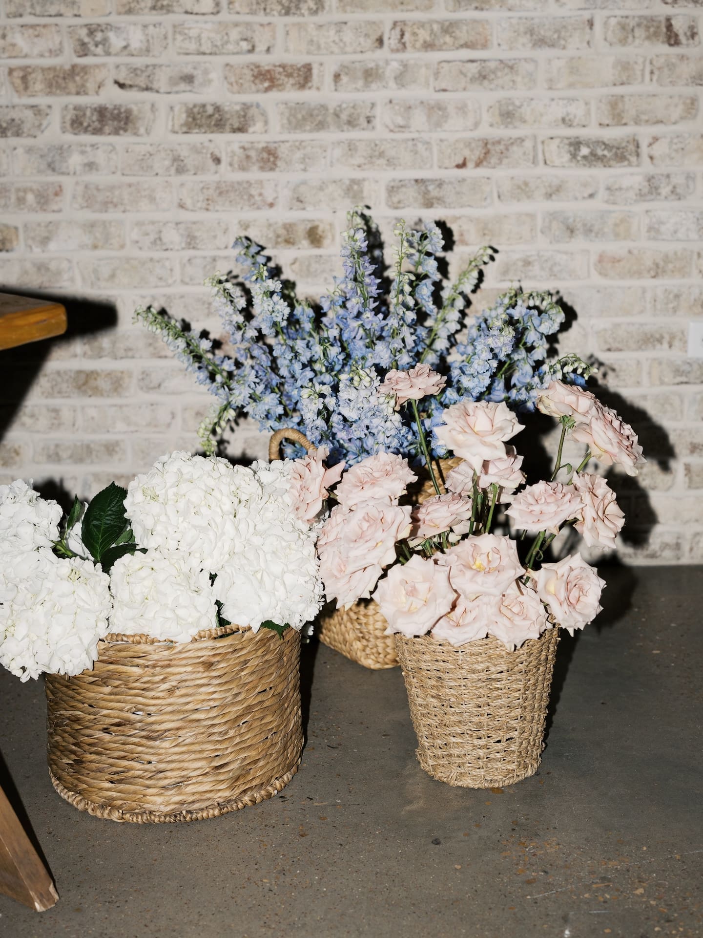 Woven baskets filled with white hydrangeas, blush roses, and blue delphinium sit against the brick wall of the Camp Hosea reception hall.