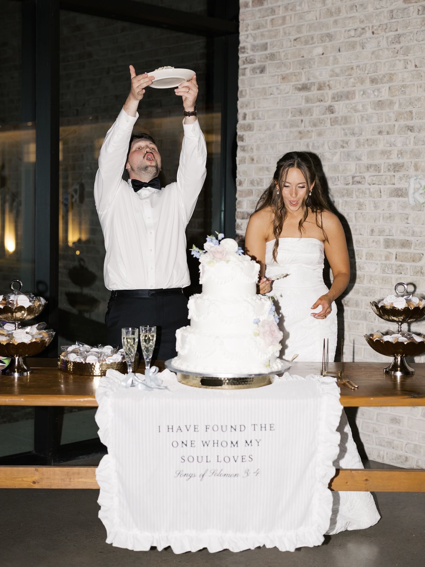 The groom dramatically tilts a plate of cake over his mouth while the bride laughs beside their tiered wedding cake, the table draped with a Song of Solomon scripture.