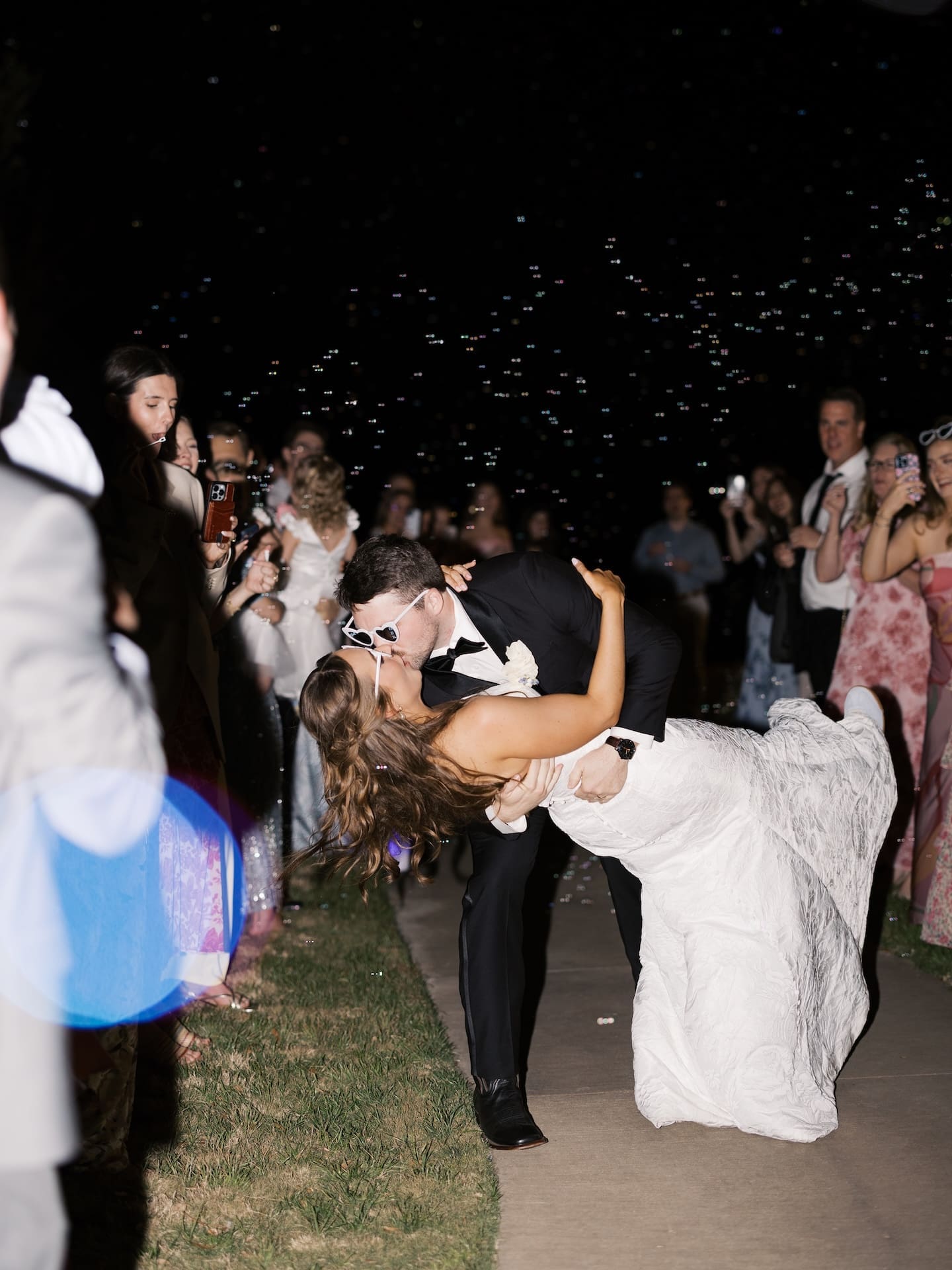 The groom dips the bride for a kiss during their bubble send-off at Camp Hosea, both in sunglasses and surrounded by cheering guests under the night sky.
