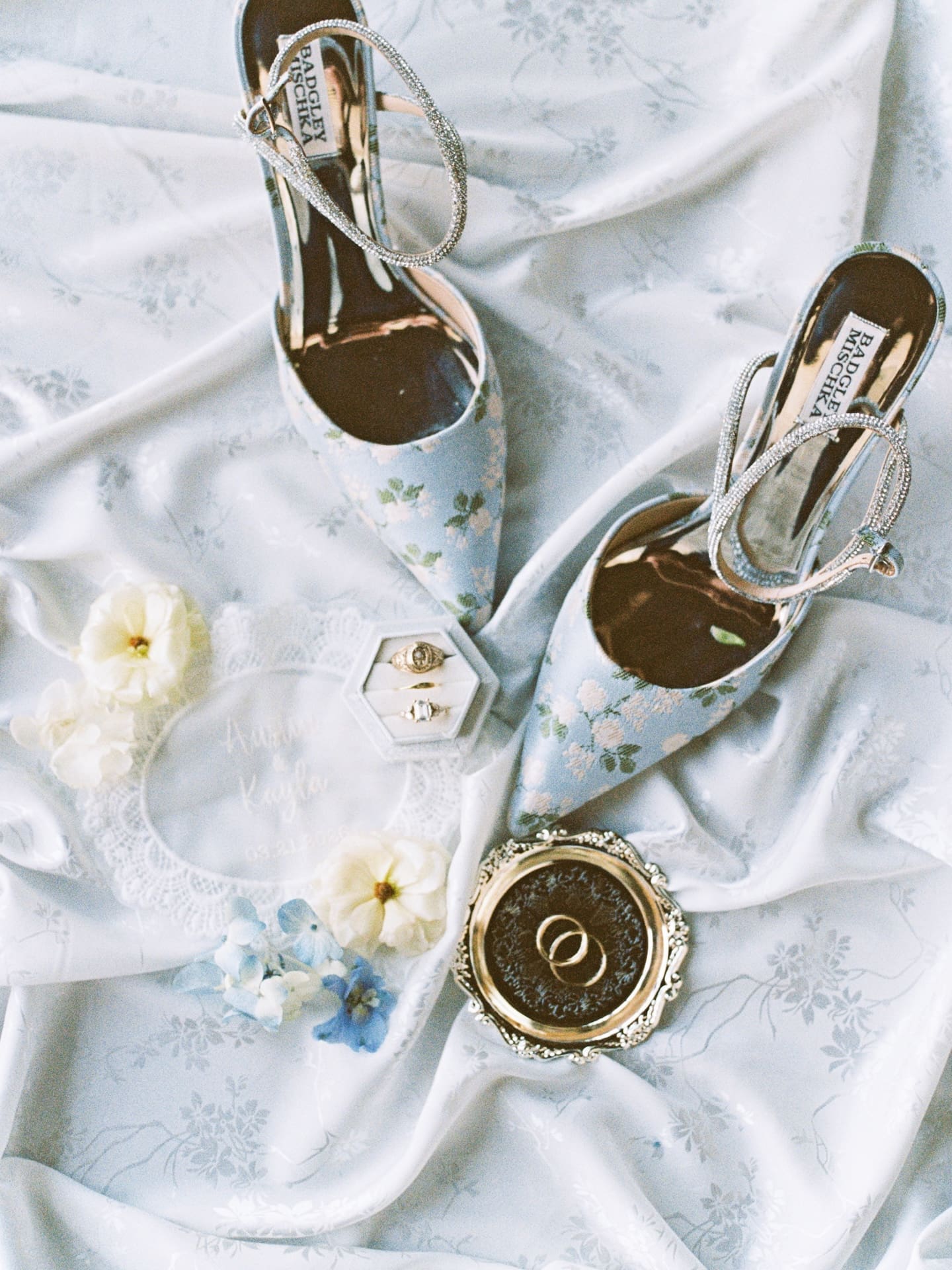 A styled flat lay of the bride's powder blue Badgley Mischka heels, wedding rings, and loose florals at Camp Hosea.