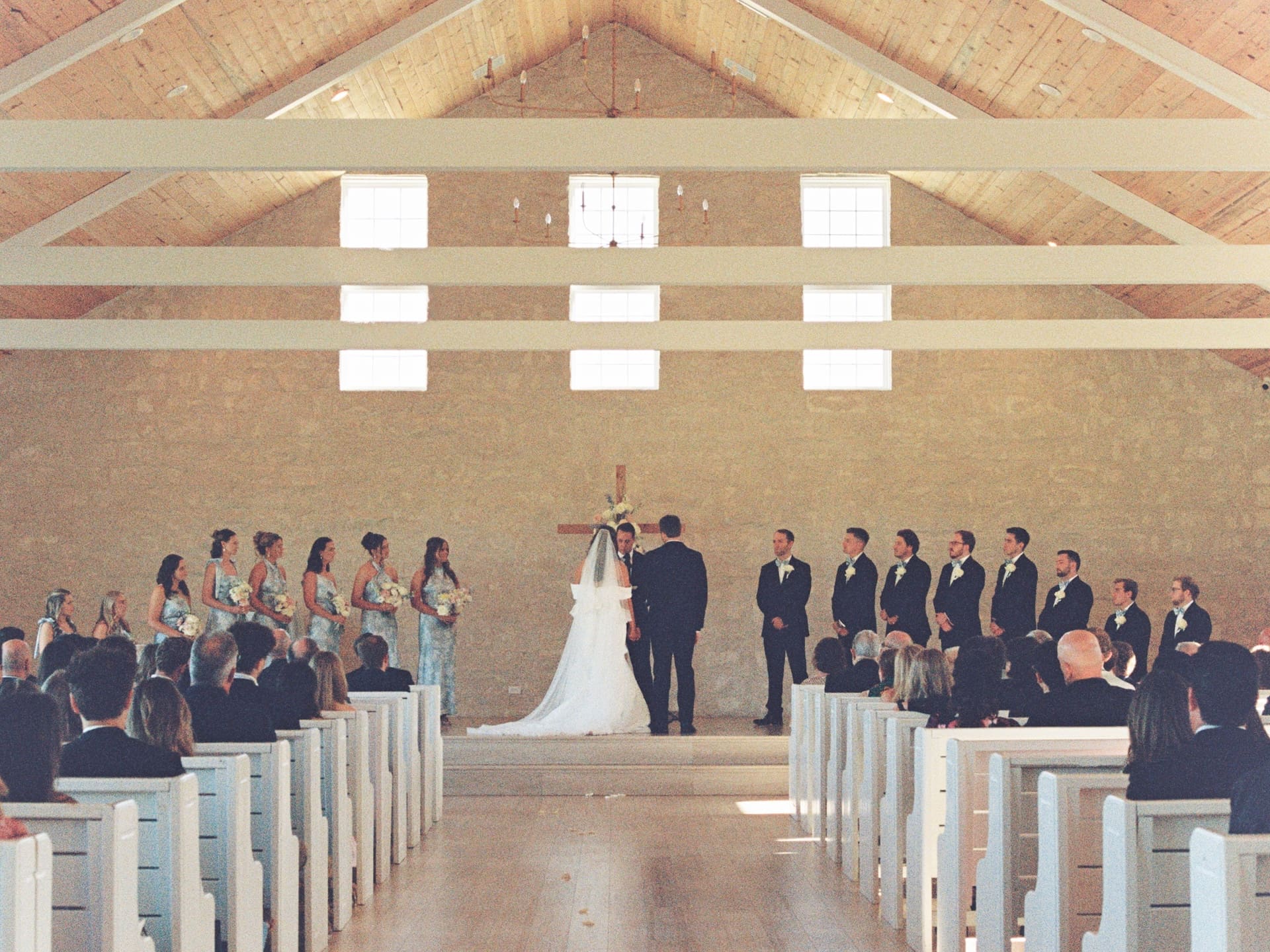 A wide film photograph of the full wedding ceremony inside the chapel at Camp Hosea, the couple at the altar surrounded by their wedding party and guests filling the white pews.