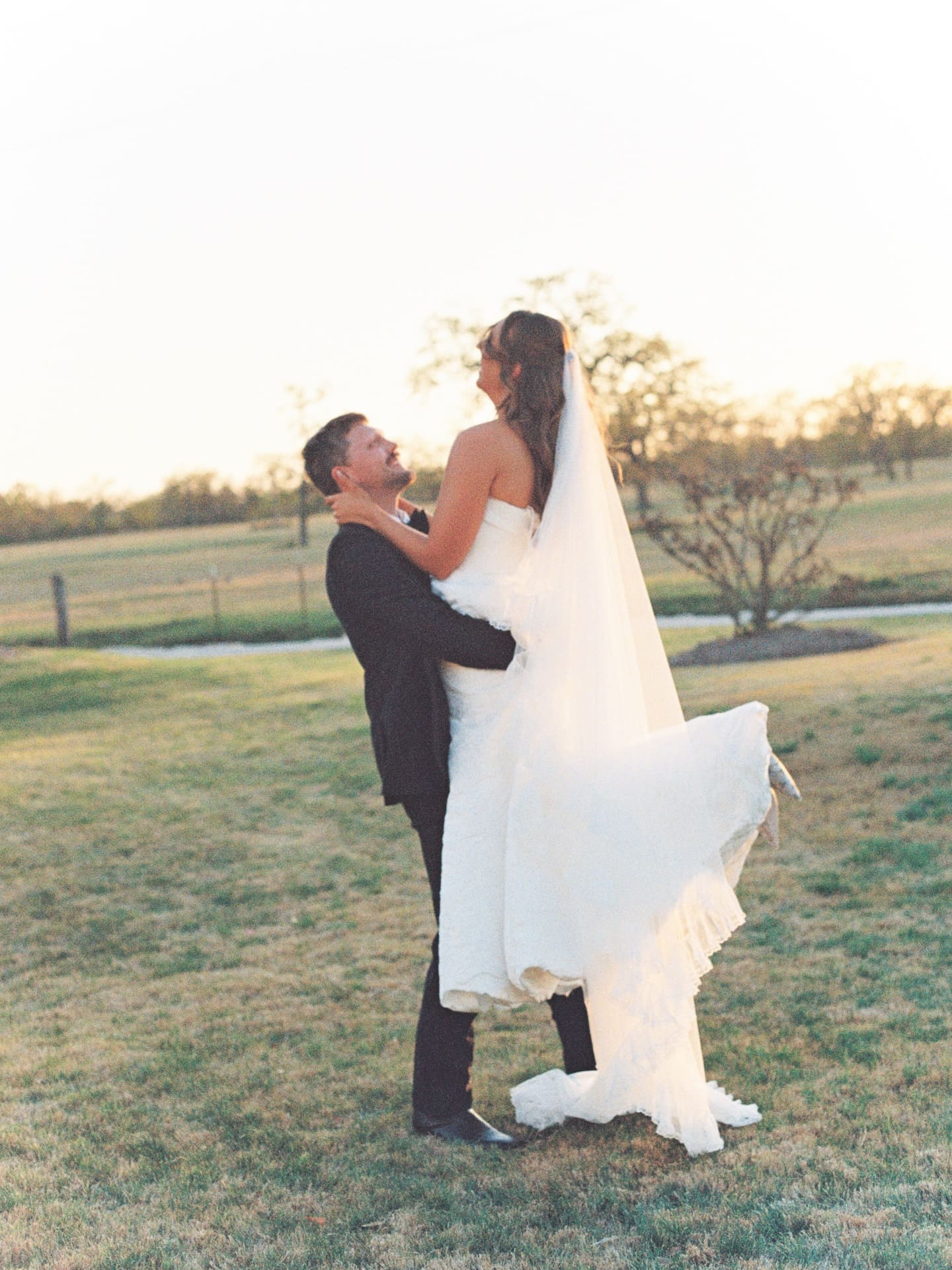 The groom lifts the bride high in the air on the golden lawn at Camp Hosea, her veil and skirt catching the last light of the setting sun.