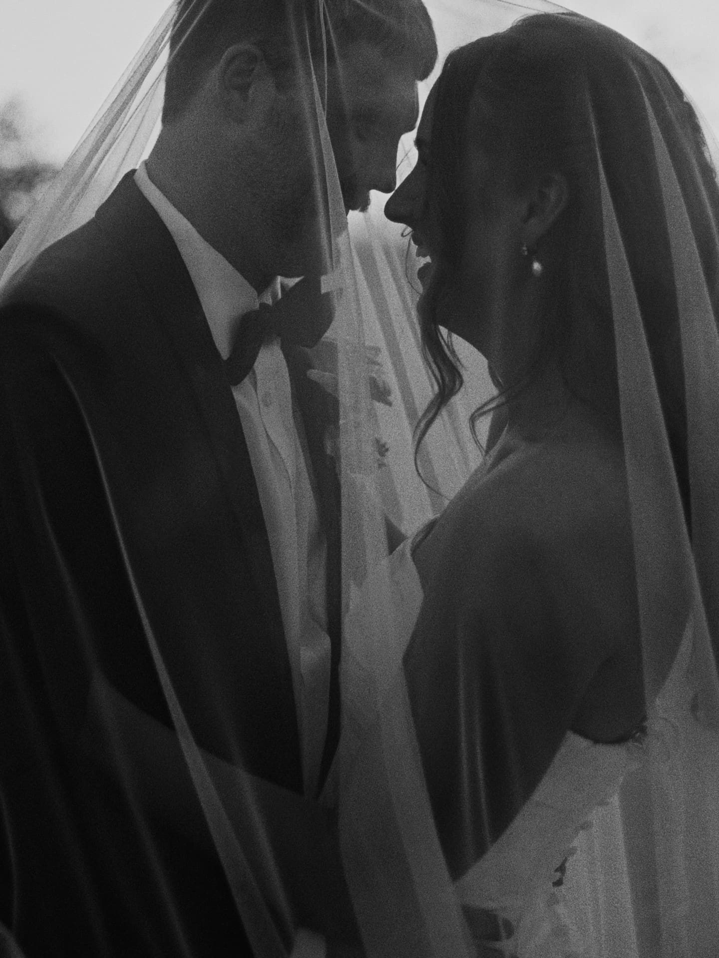 A close, intimate black and white film portrait of the bride and groom nearly nose to nose beneath her veil at dusk.