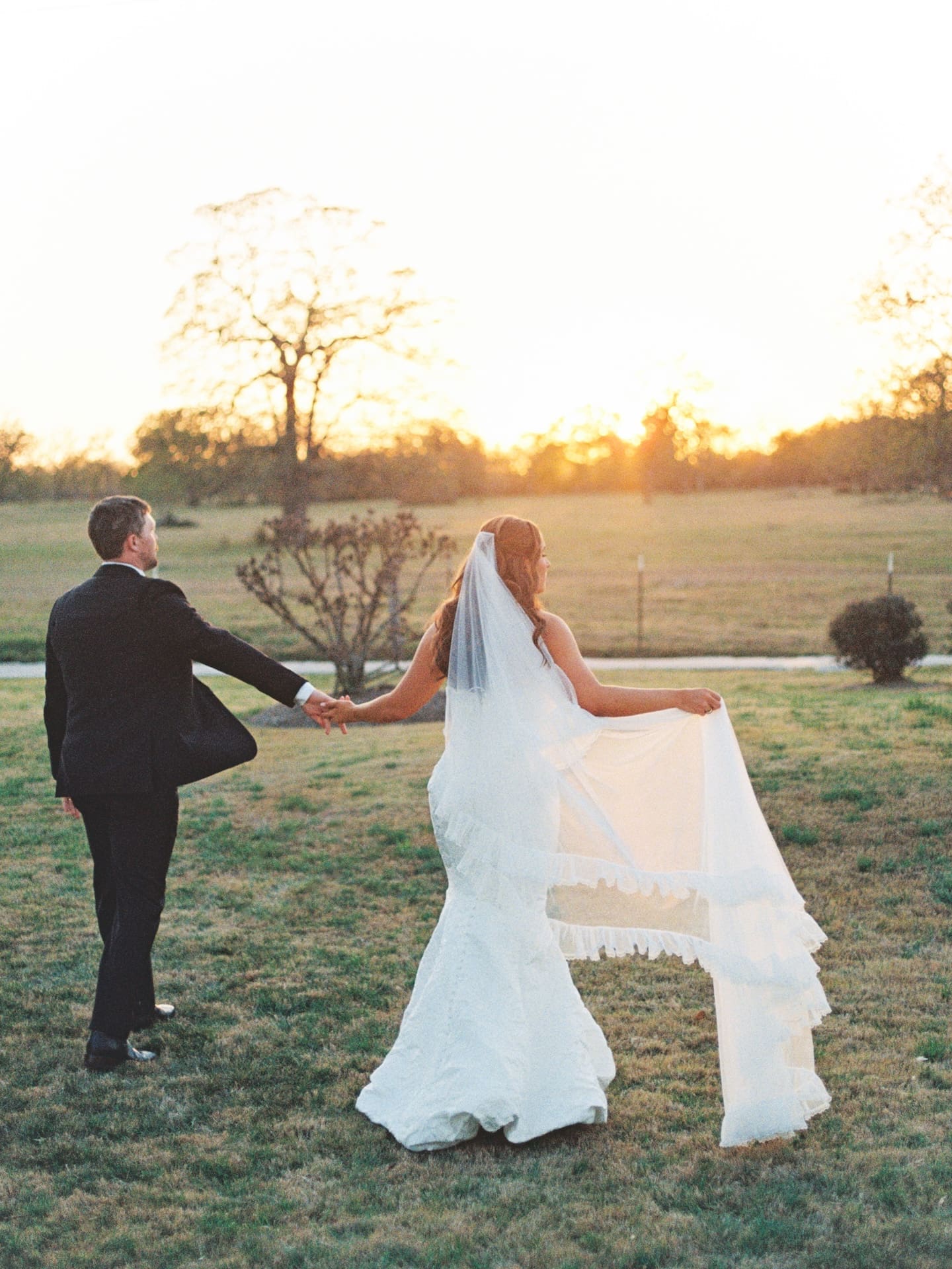 The bride and groom walk hand in hand toward a breathtaking sunset on the Camp Hosea grounds, her veil floating out behind her in the warm evening breeze.