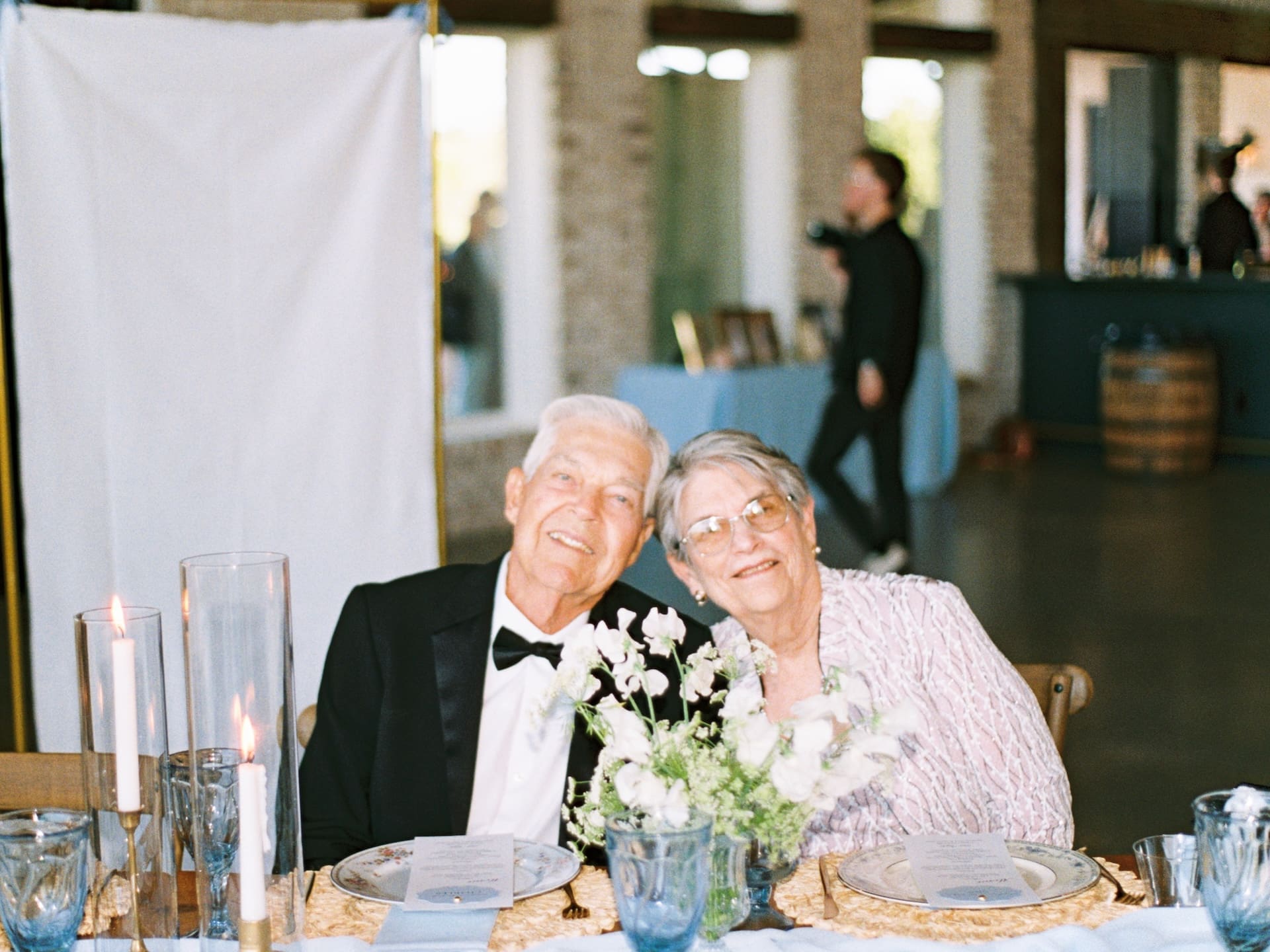 A sweet older couple leans in close at the reception table at Camp Hosea, candlelight and white florals surrounding them.