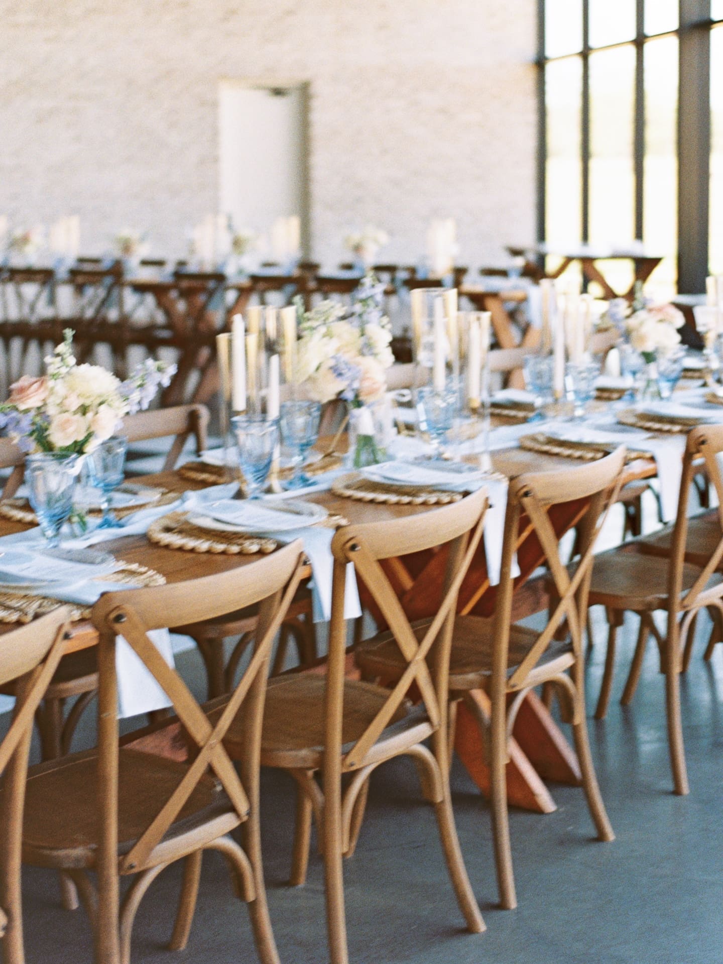 Long farm tables set for the wedding reception at Camp Hosea are lined with ivory taper candles, blue glassware, and soft blush and white florals.