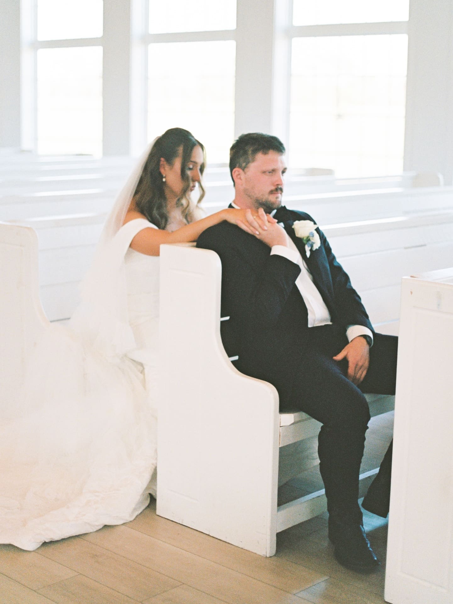 The bride and groom share a quiet, still moment together in a sunlit white chapel pew just before the ceremony.