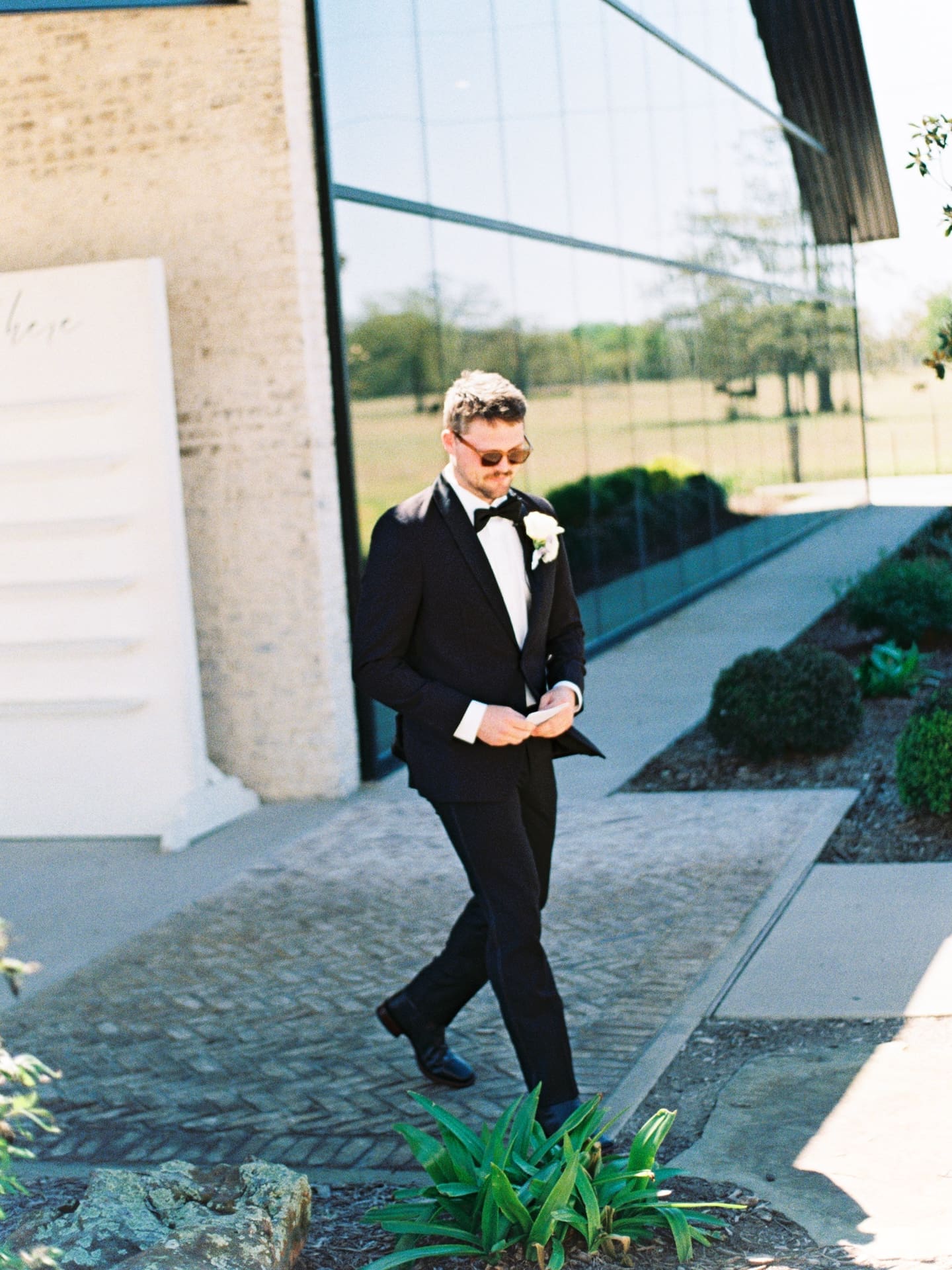 The groom walks along the glass chapel exterior at Camp Hosea in his black tuxedo, vows in hand, caught in a candid moment before the ceremony.