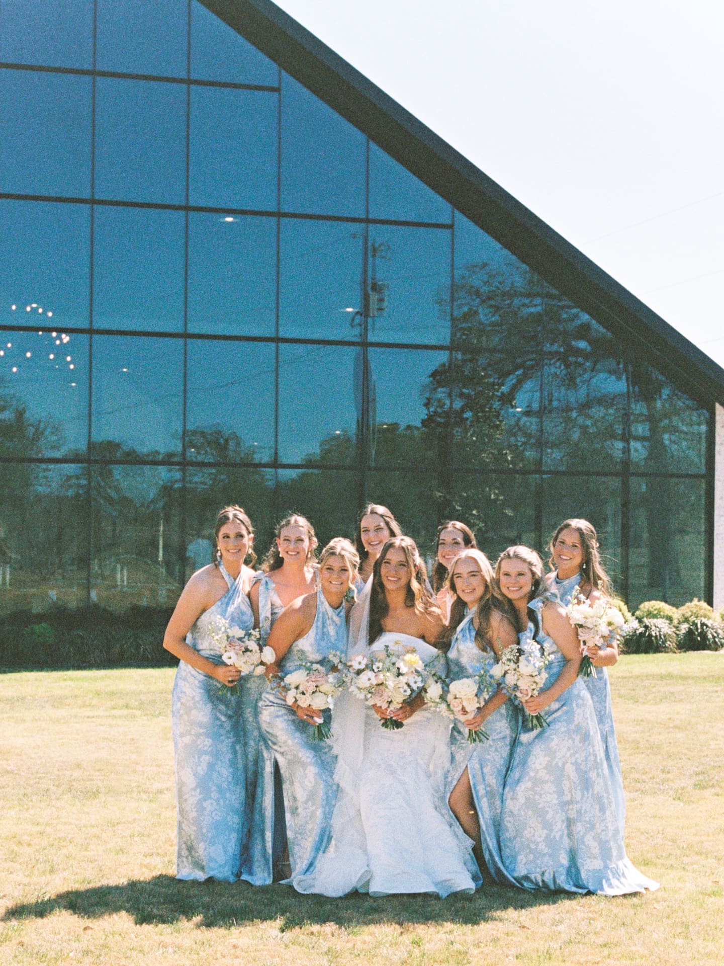 The bride and her bridesmaids in dusty blue floral gowns pose in front of the glass chapel at Camp Hosea, bouquets in hand and beaming in the afternoon sun.