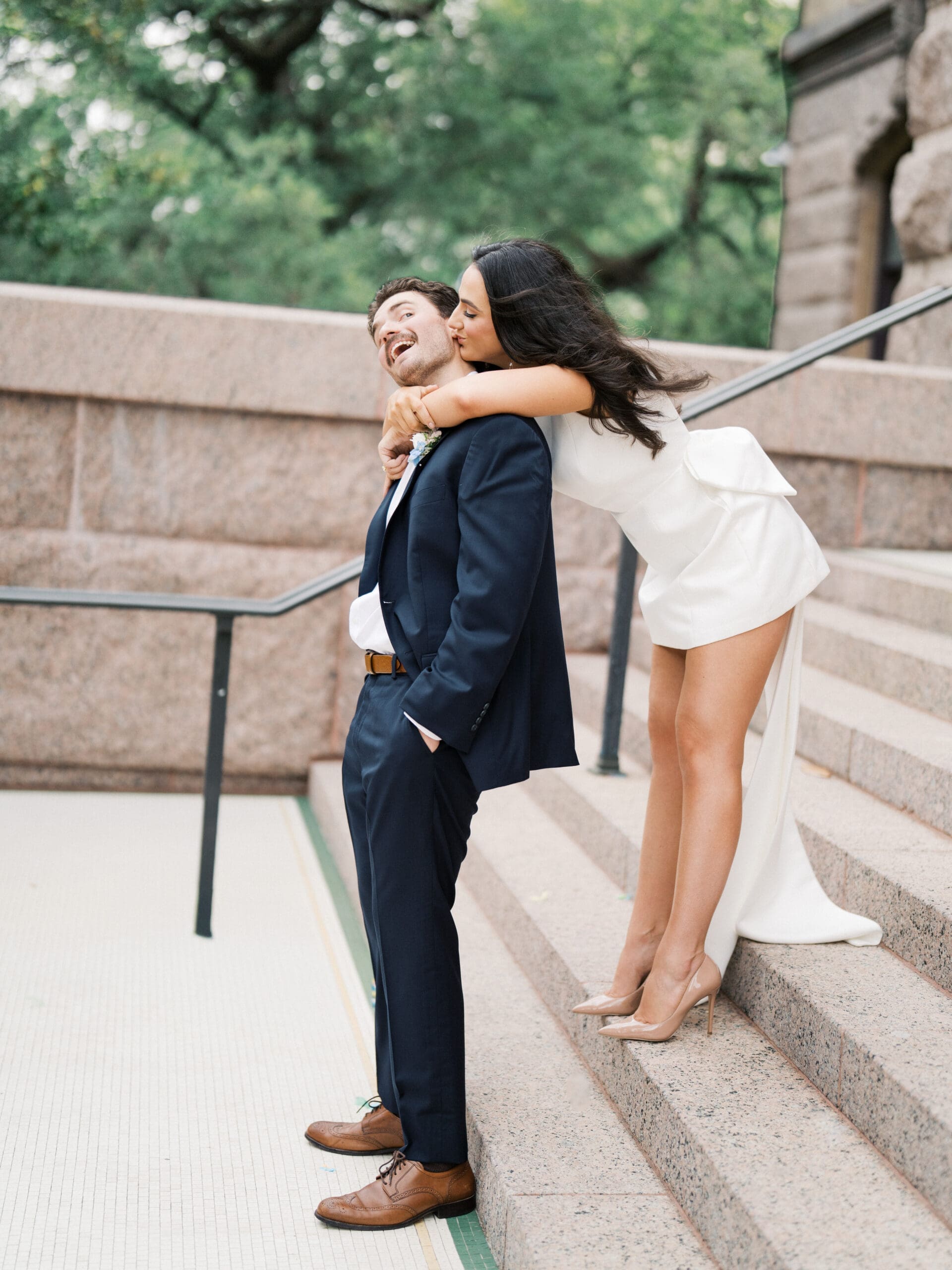 Bride hugging groom from behind on granite steps, as the bride kisses the grooms cheek over her shoulder.