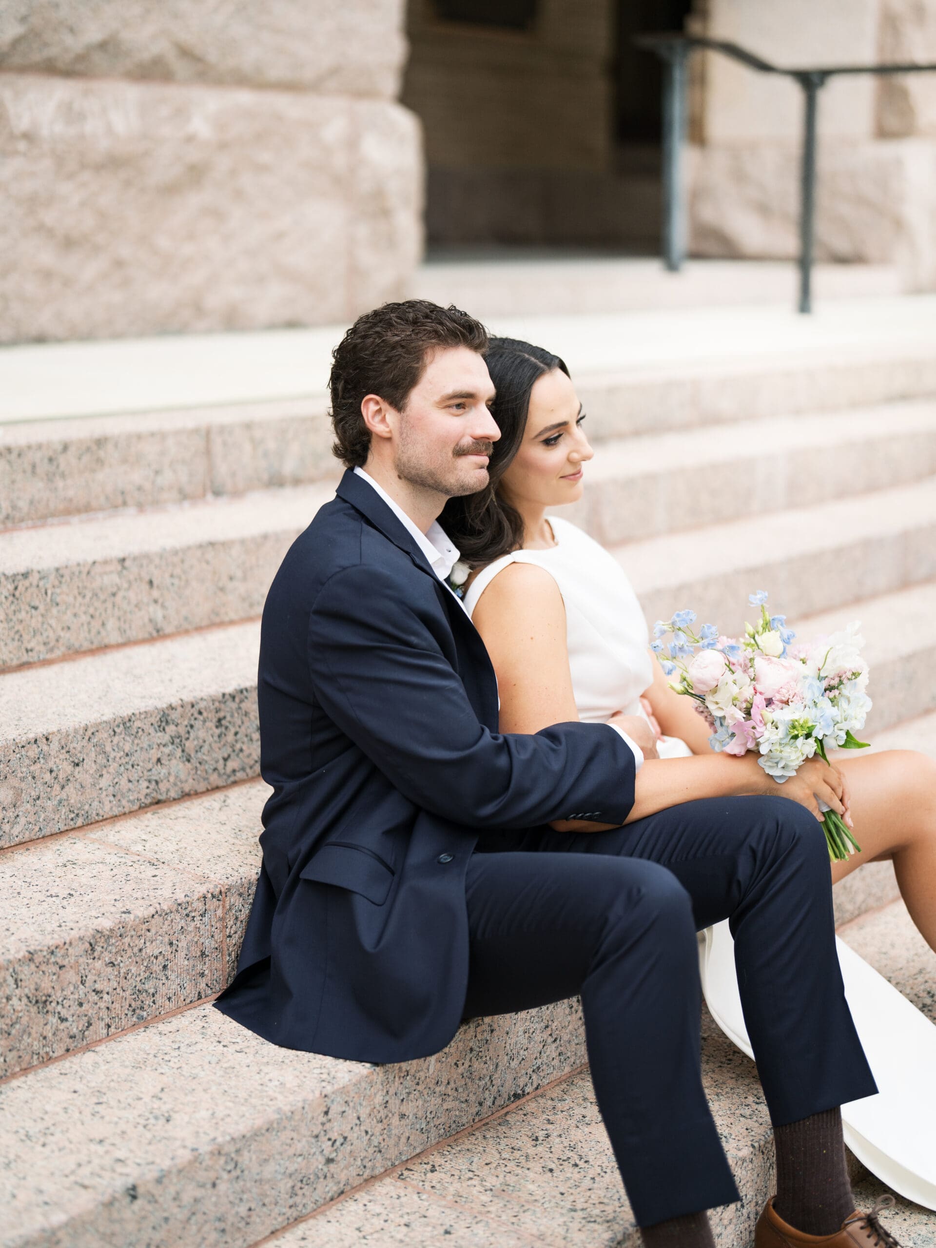 Bride and groom sitting close together on the granite steps holding a pastel bouquet
