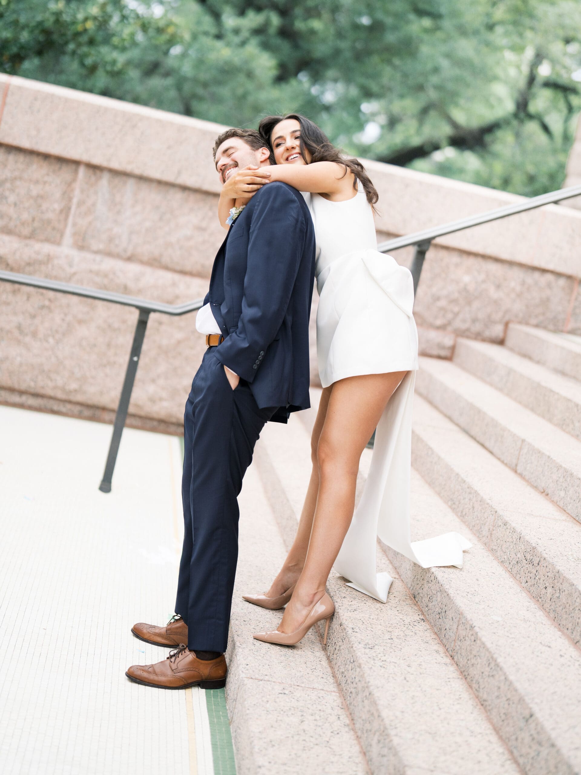 Bride hugging groom from behind on granite steps, both laughing
