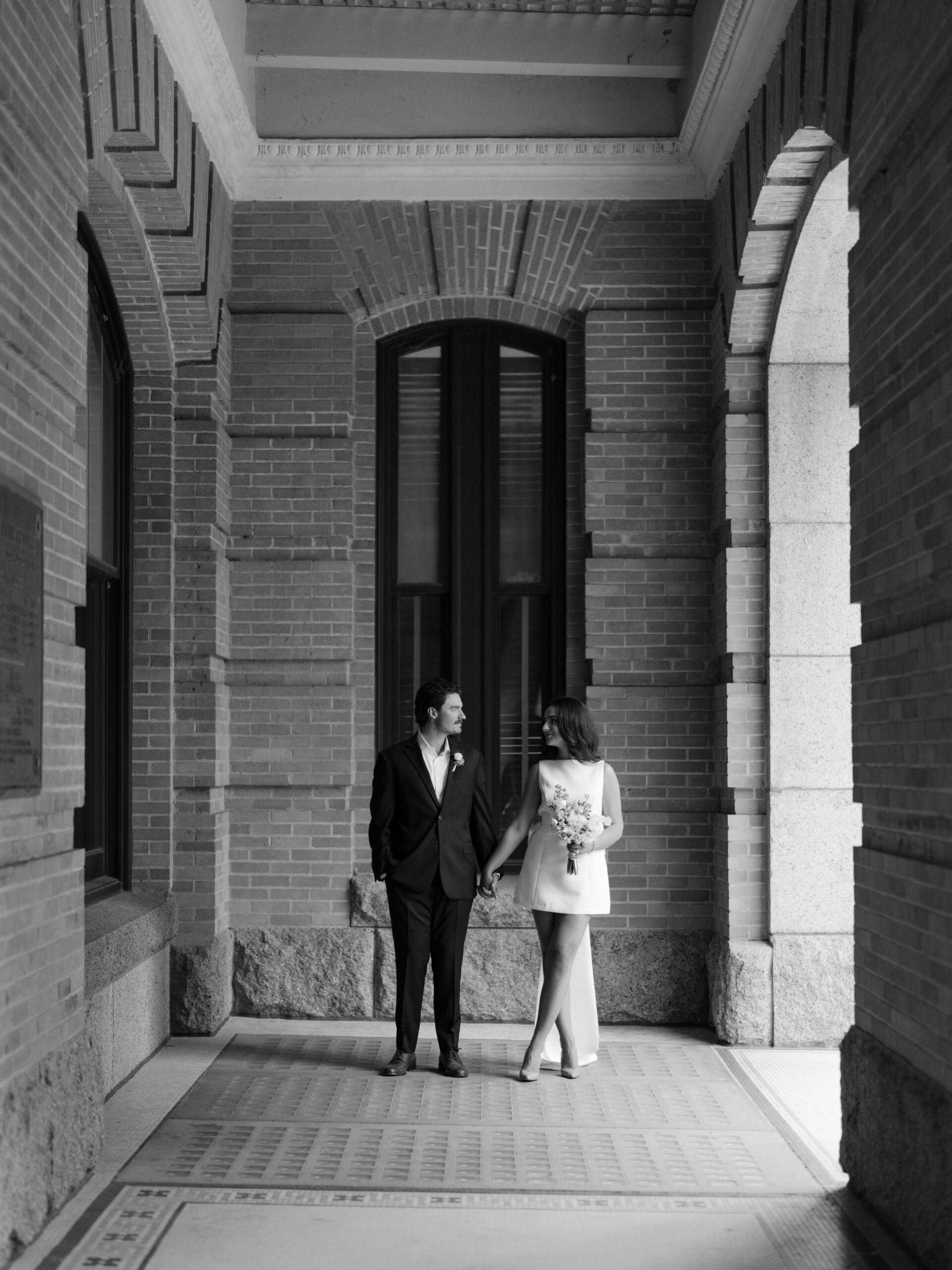 Black and white portrait of the couple holding hands in a brick archway corridor
