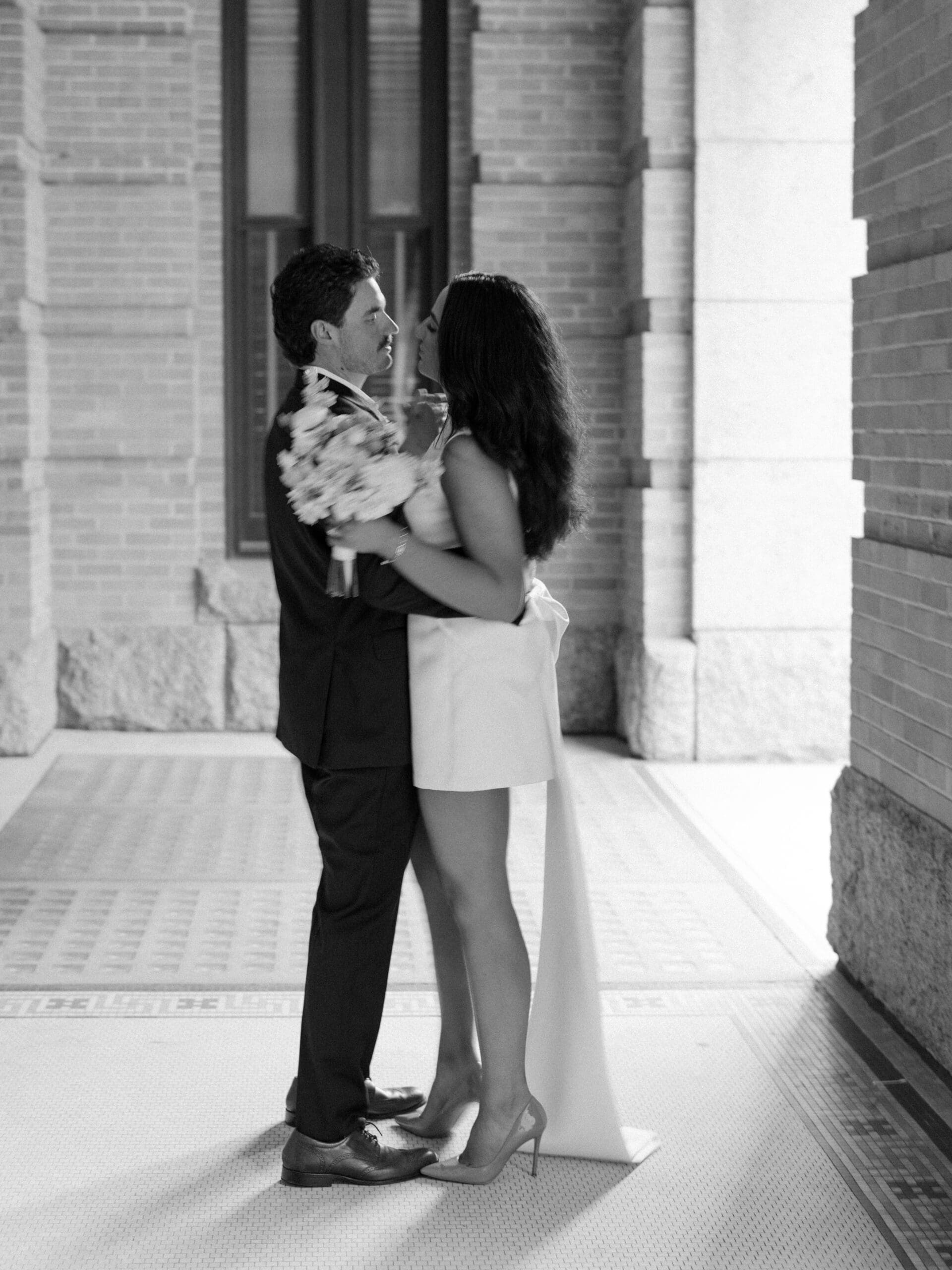 Black and white photo of the couple embracing and gazing at each other in a sun-lit courthouse corridor
