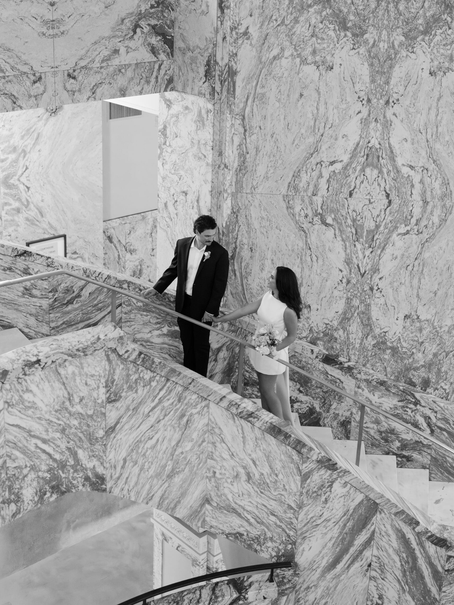 Black and white overhead shot of the couple descending a sweeping marble staircase

