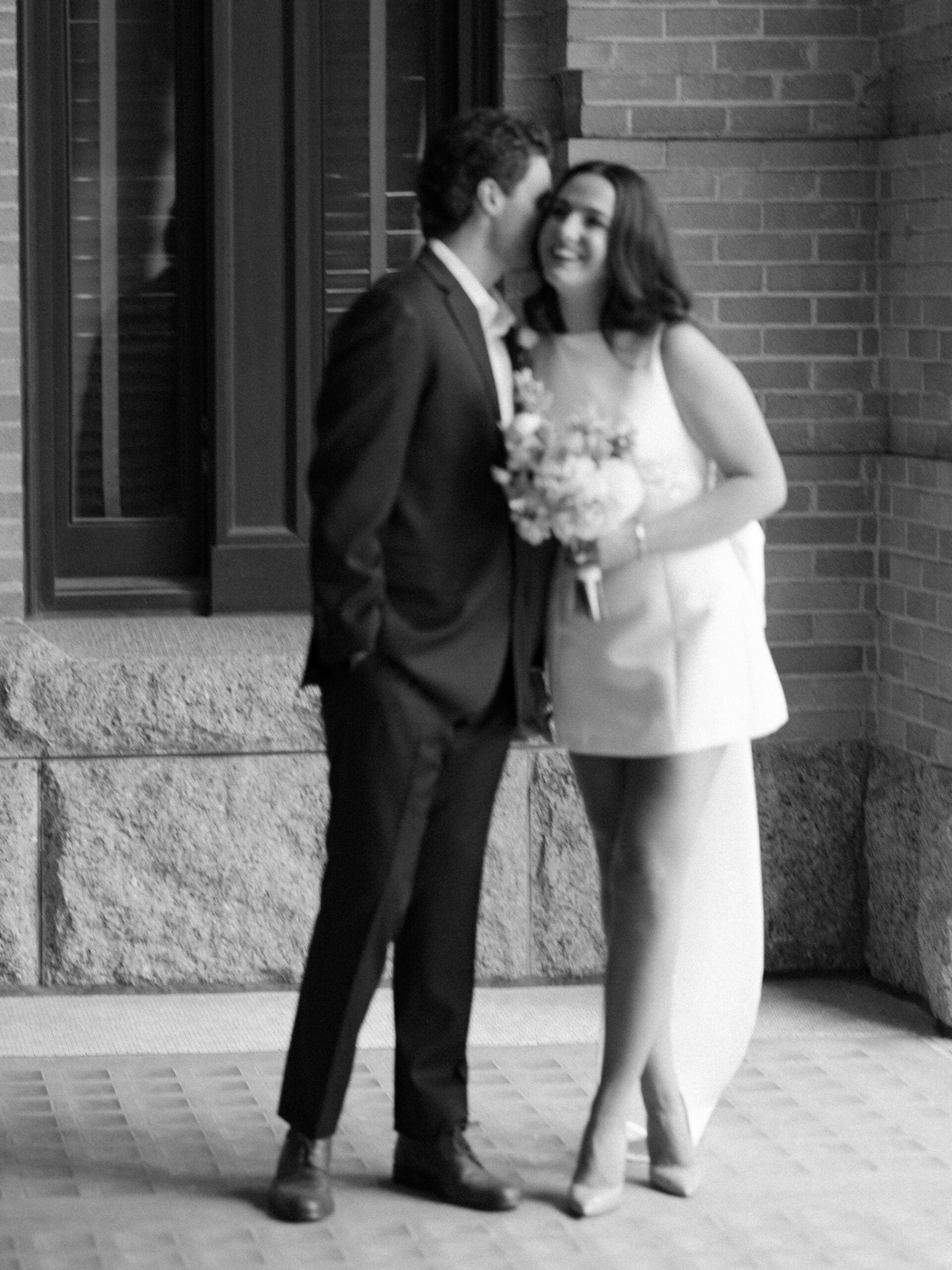 Groom whispering in his bride's ear outside a historic brick building
