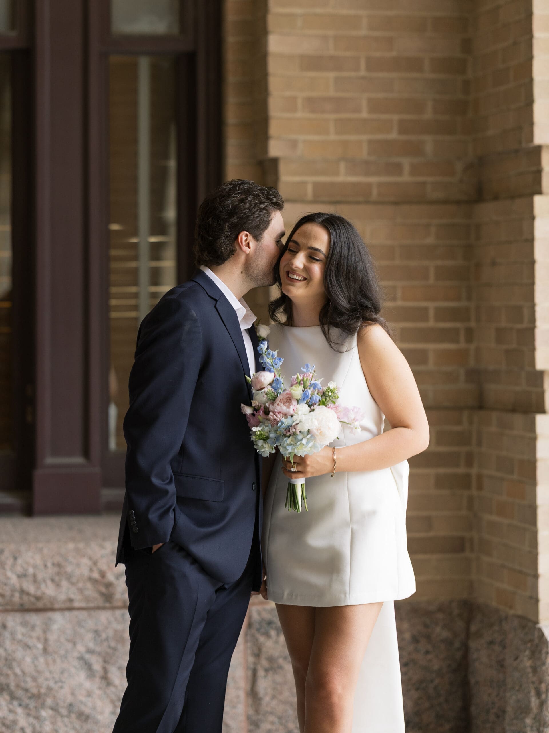 Groom kissing his bride on the cheek as she laughs outside the historic brick courthouse
