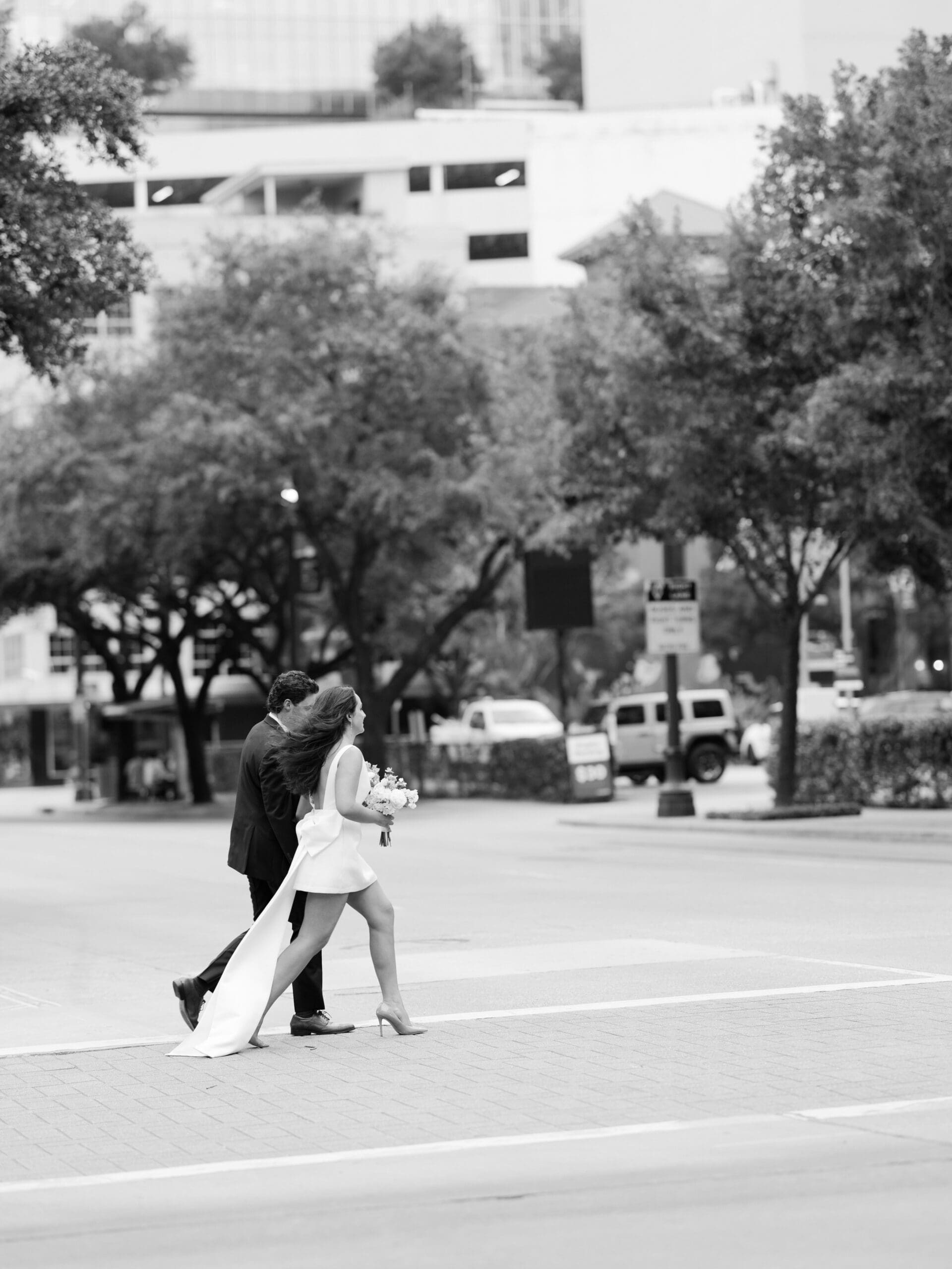 A bride and groom hold hands while crossing a city street in black and white

