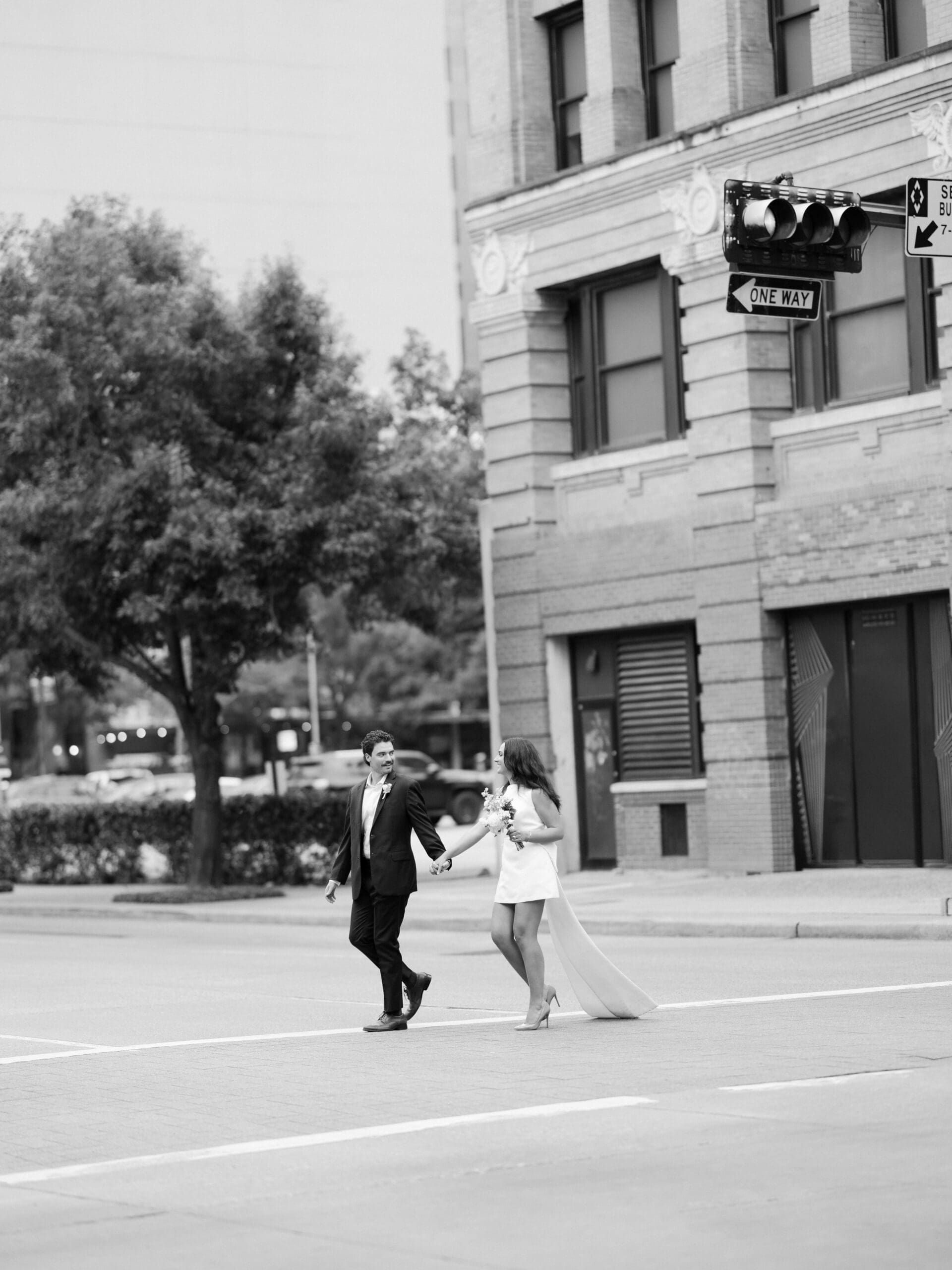 A bride and groom hold hands while crossing a city street in black and white
