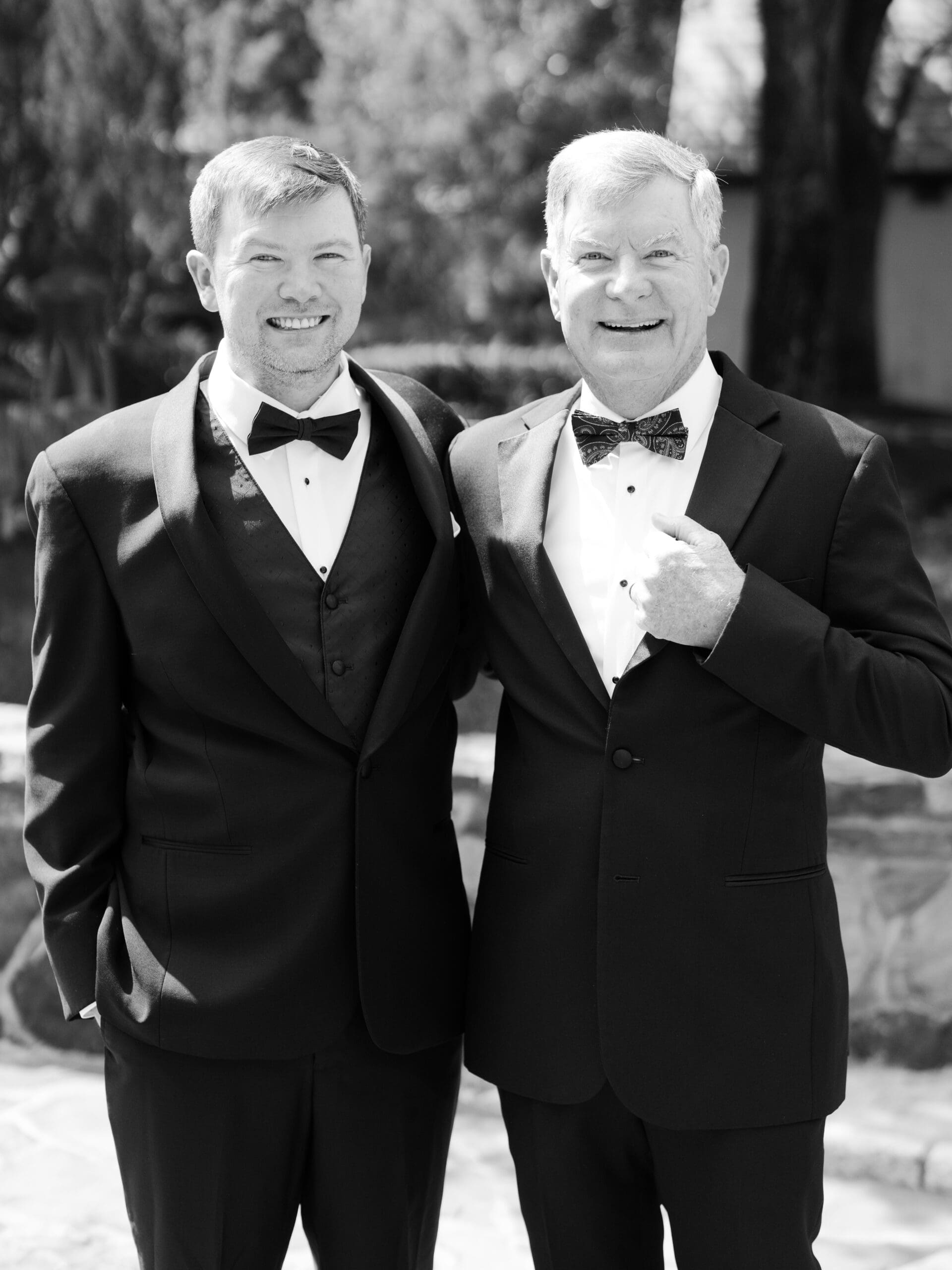 Black and white portrait of a groom and his father smiling together outdoors in matching black tuxedos.
