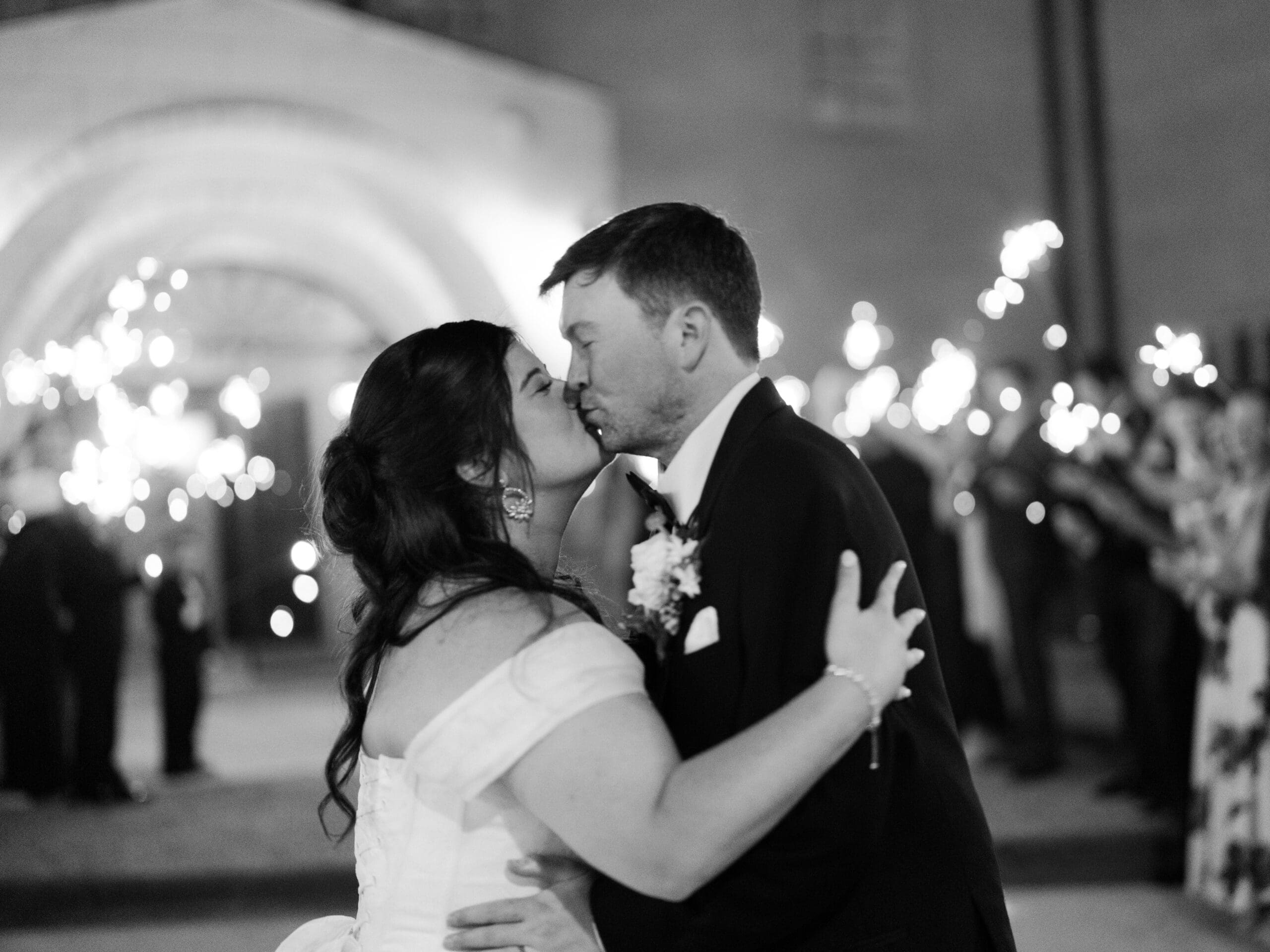 Black and white image of a bride and groom sharing a kiss during their sparkler send-off outside The Lanier Theological Library, with bokeh lights glowing all around them.
